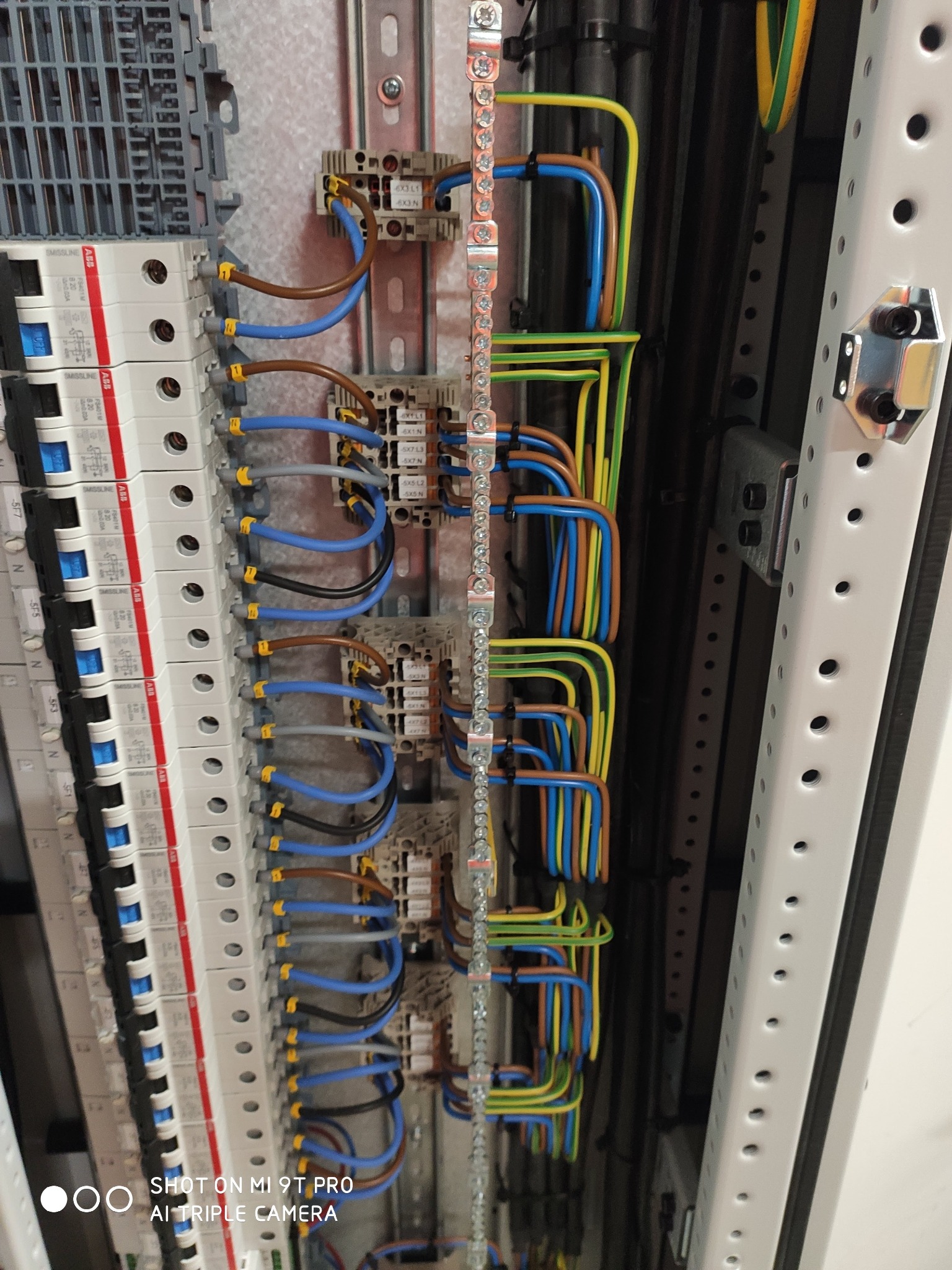 Panel elektryczny z bezpiecznikami i okablowaniem w kolorach: niebieskim, brązowym, żółto-zielonym. Widoczne złącza i szyny DIN. Element instalacji elektrycznej w rozdzielnicy.