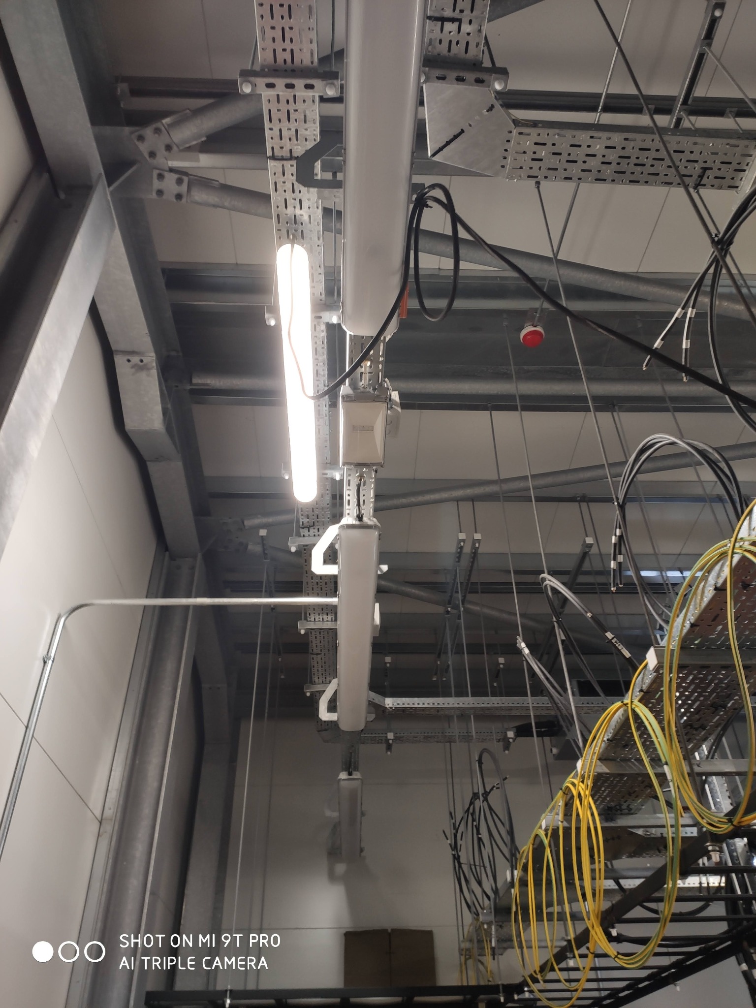 Biała lampa LED zamontowana na suficie w hali przemysłowej, widoczne metalowe korytka kablowe, liczne kable i rury. Oświetlenie sufitowe w nowoczesnym stylu.