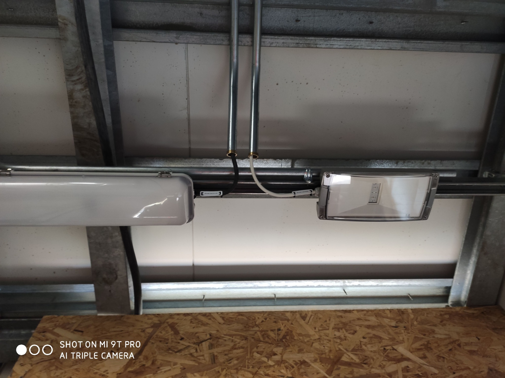 Dwa różne typy lamp sufitowych zamontowane na metalowych rurach w pomieszczeniu z płytami OSB. Widoczne przewody elektryczne i konstrukcja sufitu.