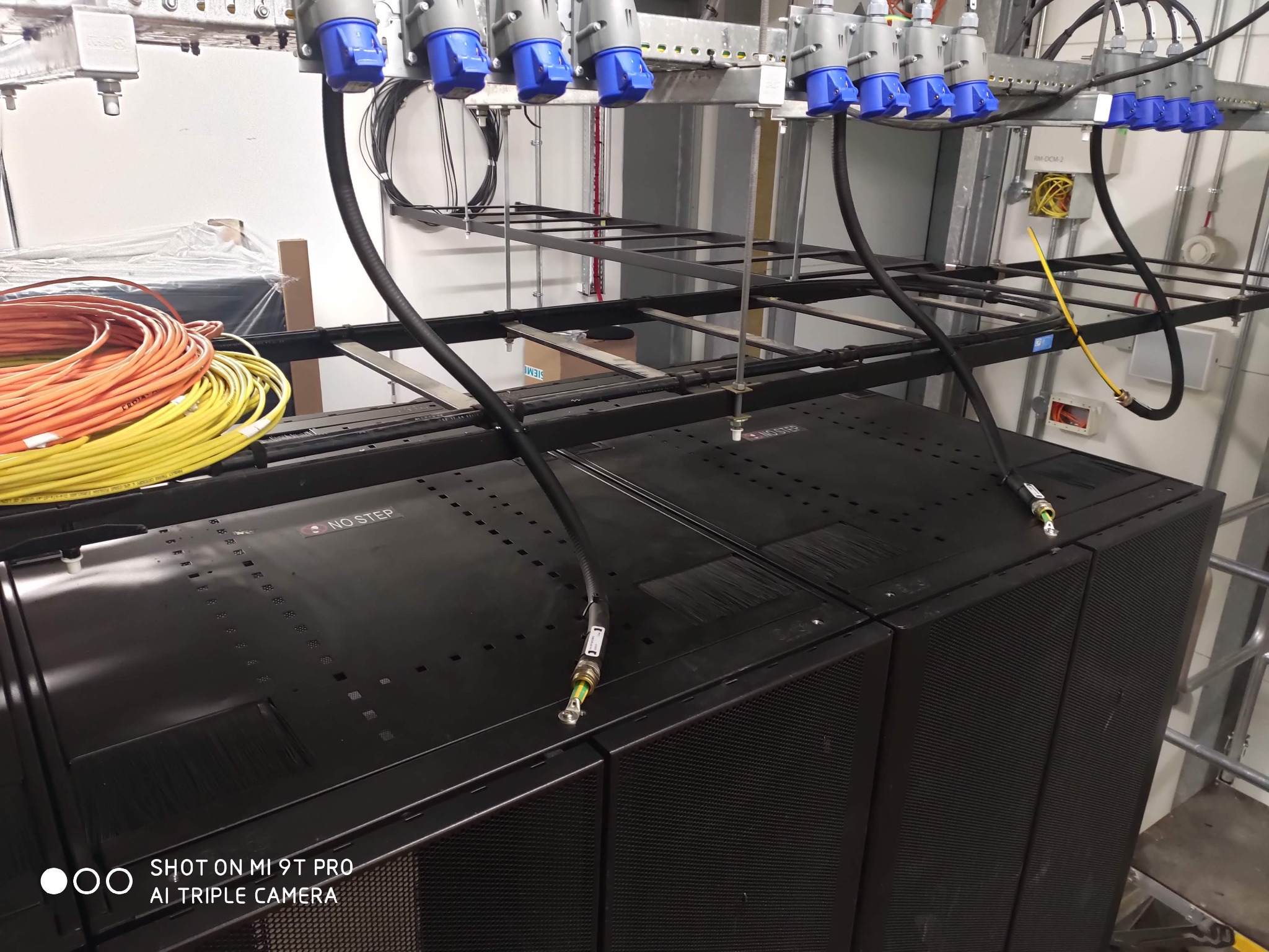 Instalacja elektryczna w serwerowni: kable z niebieskimi wtyczkami, czarne szafy rack z napisem 'NO STEP' i pomarańczowe kable. Widoczne metalowe konstrukcje pod sufitem.