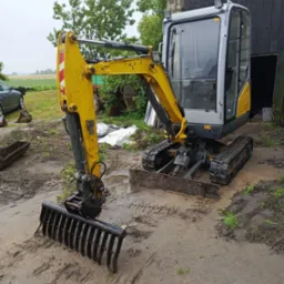 Żółto-szara mini koparka gąsienicowa z zębami do niwelacji terenu stoi na błotnistym placu budowy. Widoczny budynek gospodarczy w tle.