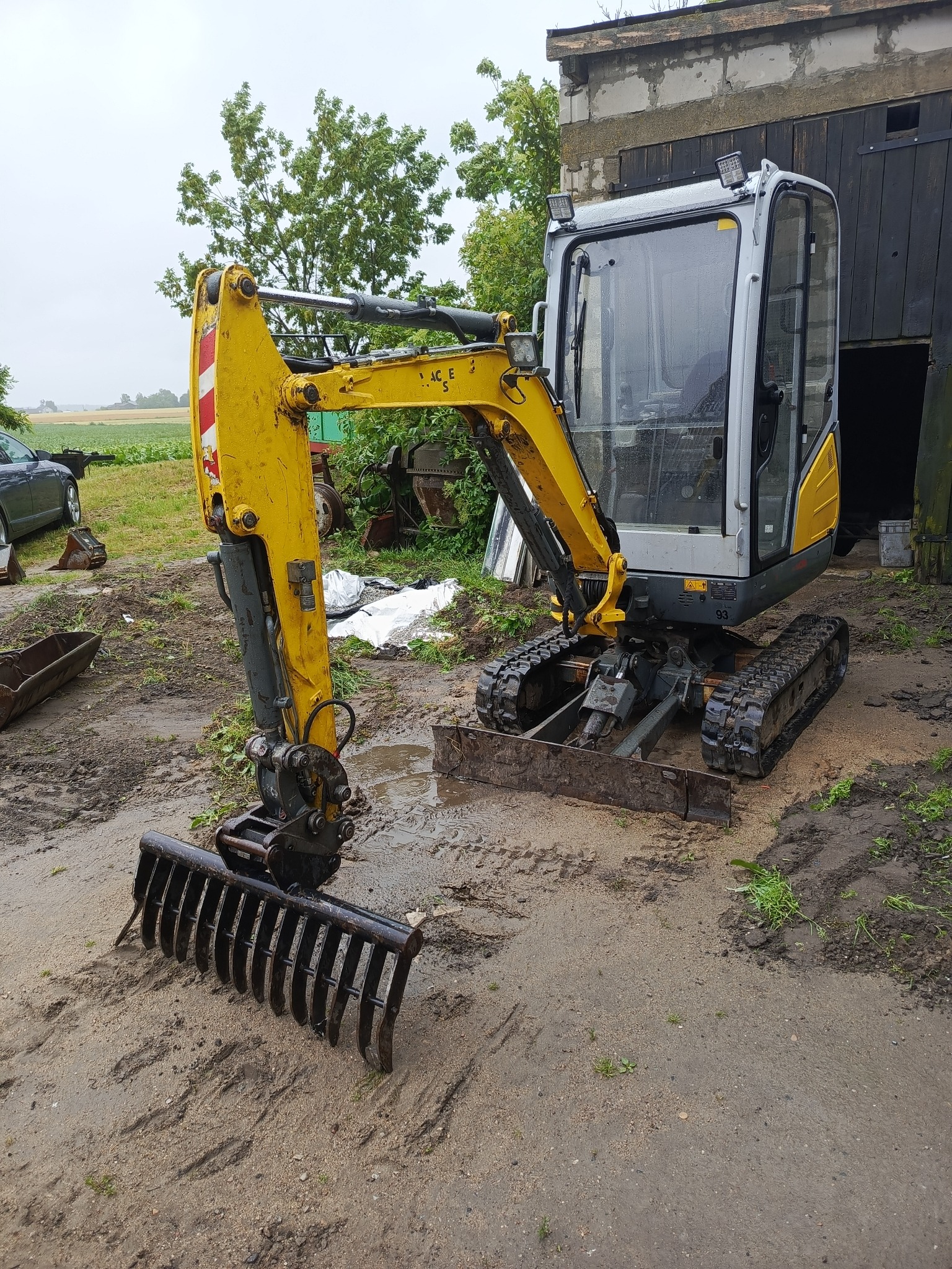 Żółto-szara mini koparka gąsienicowa z zębami do niwelacji terenu stoi na błotnistym placu budowy. Widoczny budynek gospodarczy w tle.