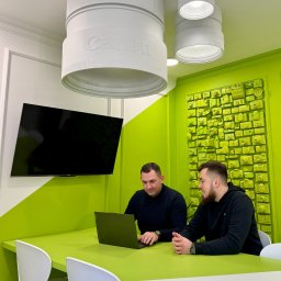 Up Blue Sp. z o. o. - Dwóch mężczyzn pracuje przy zielonym stole w biurze z dekoracjami nawiązującymi do fotografii, w tym lampy w kształcie obiektywów Canon.