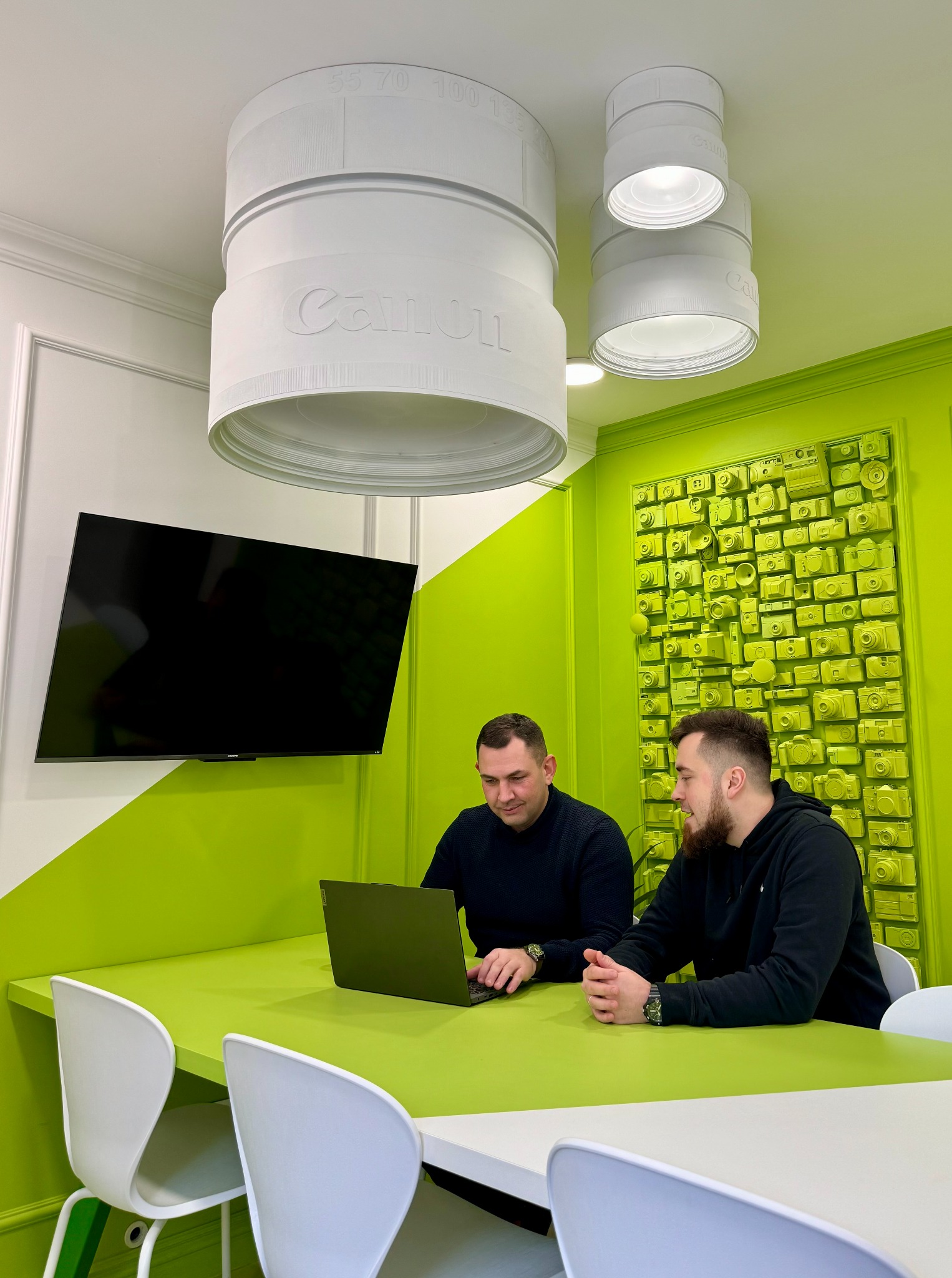 Dwóch mężczyzn pracuje przy zielonym stole w biurze z dekoracjami nawiązującymi do fotografii, w tym lampy w kształcie obiektywów Canon.