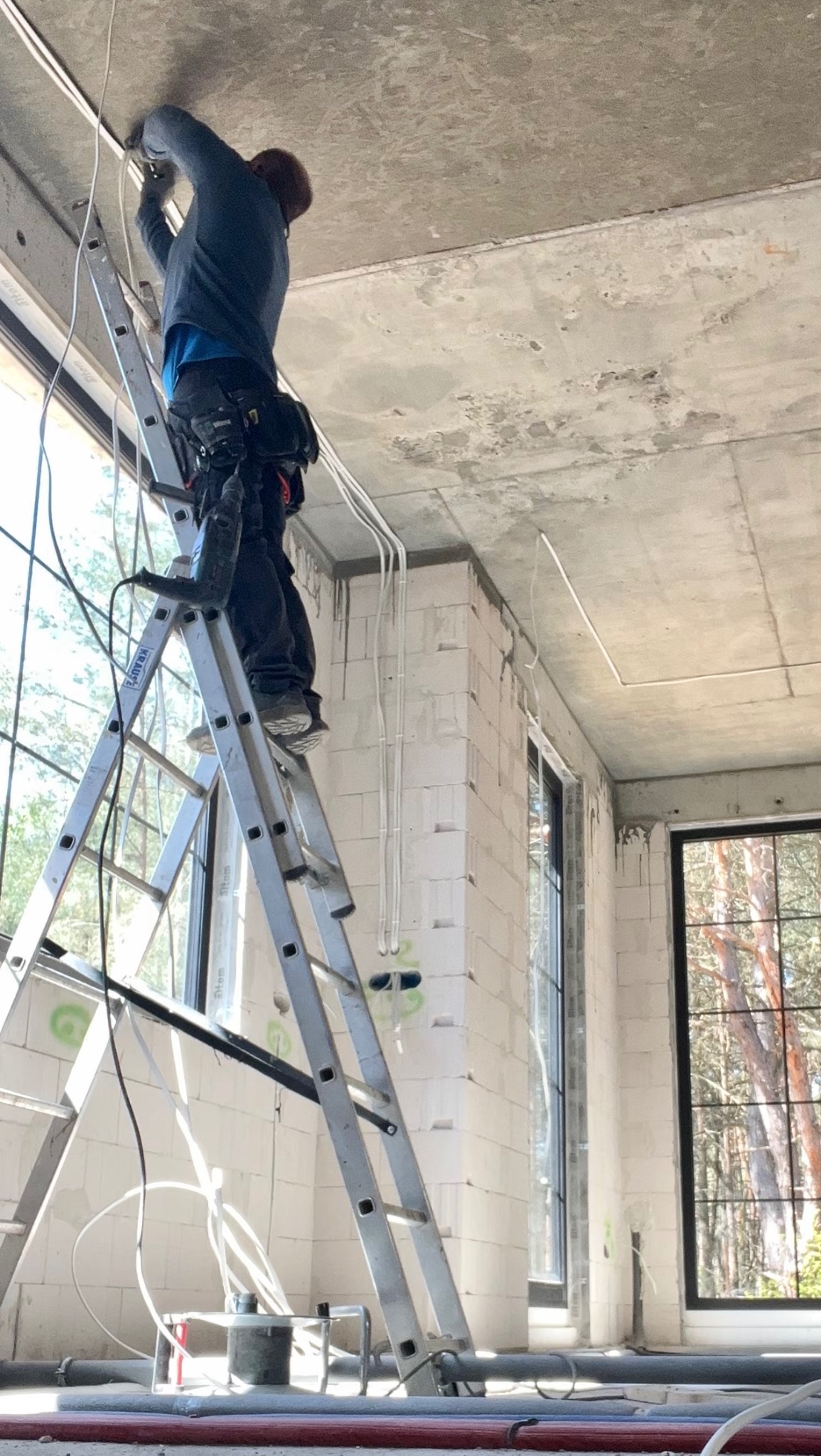Elektryk na drabinie montuje białe kable na suficie w surowym wnętrzu. Widoczne okna z widokiem na las i narzędzia pracy. Instalacja elektryczna w trakcie realizacji.