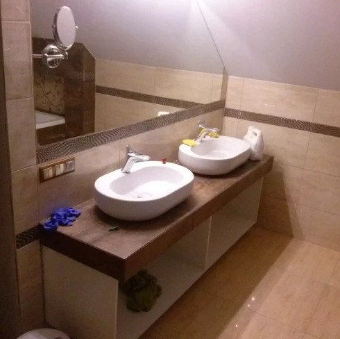 Wykończona łazienka z dwiema owalnymi umywalkami nablatowymi, chromowanymi bateriami i lustrem na ścianie z beżowych płytek. Widoczne niebieskie rękawice robocze.