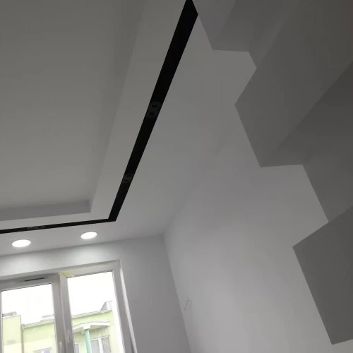 Białe wnętrze z podwieszanym sufitem z czarnymi akcentami i oświetleniem punktowym, widok na okno i fragment schodów.