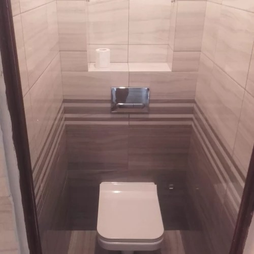 Kompaktowa toaleta w wykończonej łazience z beżowo-brązowymi płytkami, wnęką na papier toaletowy i chromowanym przyciskiem spłukiwania.