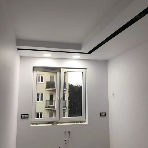 Białe wnętrze po remoncie z oknem, widok na blok mieszkalny. Czarny pas sufitowy z oświetleniem. Gładkie ściany, gniazdka elektryczne po obu stronach okna.