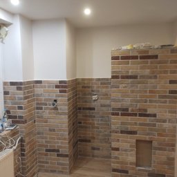 Leszek Zieleniewski - Częściowo wykończona kuchnia z ceglaną ścianą do połowy wysokości i drewnianą podłogą. Widoczne gniazdka elektryczne i fragment grzejnika.