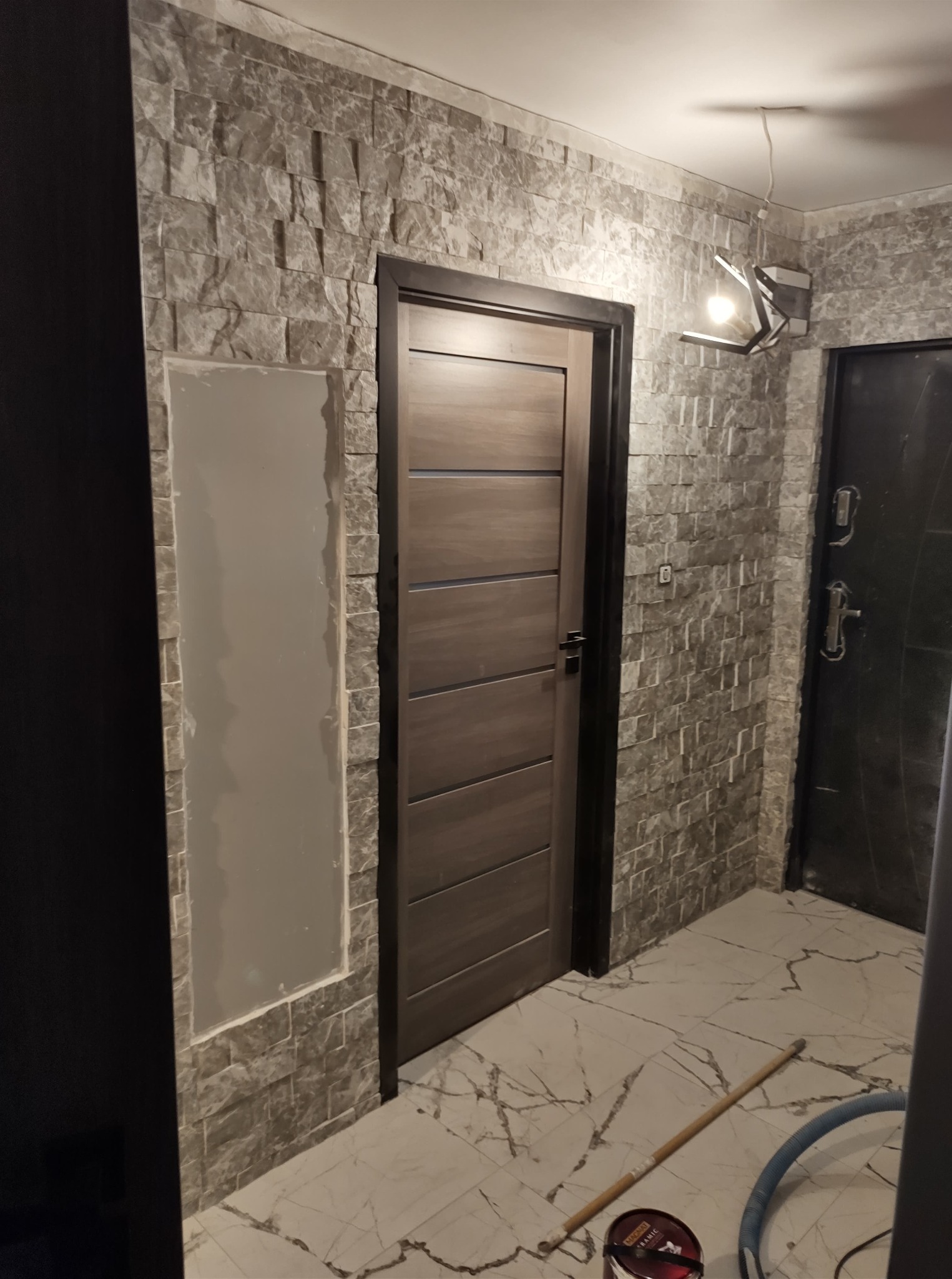 Wnętrze po remoncie: kamienna okładzina ściany, drewniane drzwi z czarną ościeżnicą. Widoczny fragment płyty karton-gips i narzędzia na podłodze.