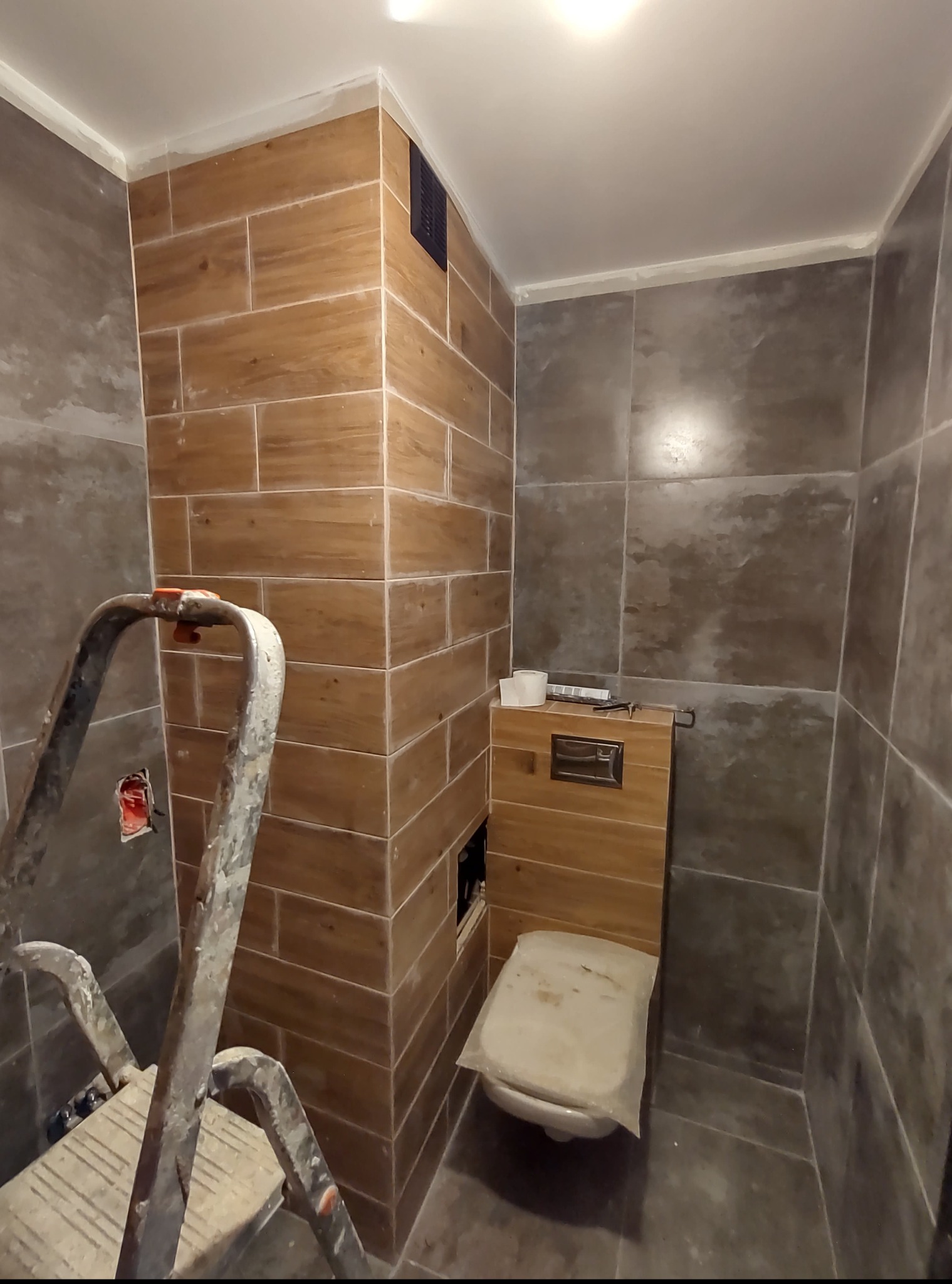 Remont łazienki: brązowe i szare płytki imitujące drewno, częściowo zakryta toaleta, drabina malarska w rogu. Widoczne akcesoria i narzędzia.