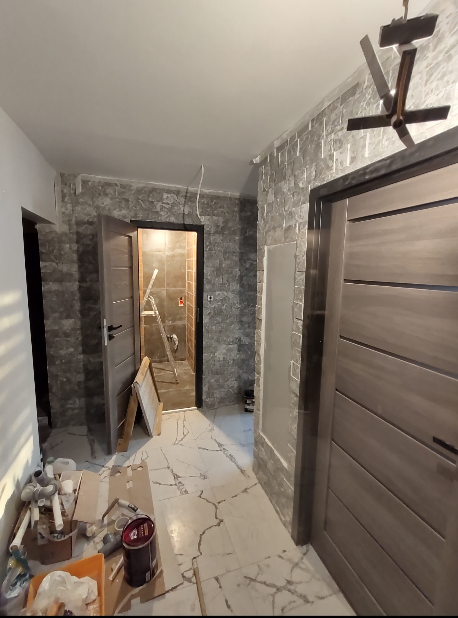 Wnętrze w trakcie wykończenia: marmurowe płytki podłogowe, ściana z dekoracyjną okładziną, otwarte drzwi do łazienki z drabiną, widoczne narzędzia i materiały budowlane.