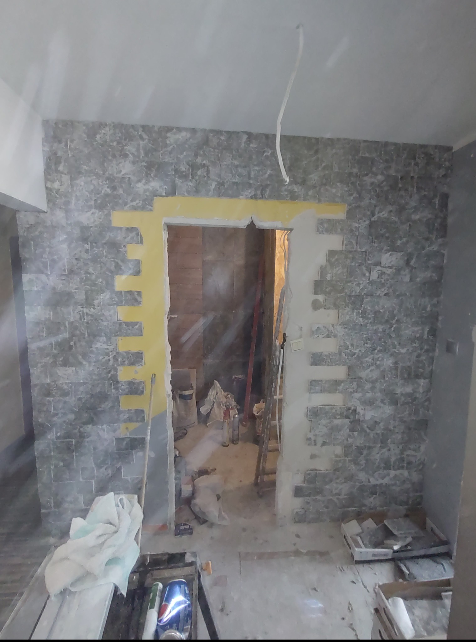 Montaż dekoracyjnej okładziny ściennej imitującej kamień w przedpokoju, z żółtymi akcentami wokół otworu drzwiowego. Widoczne narzędzia i materiały budowlane.