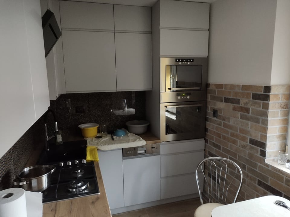 Nowoczesna kuchnia z szarymi szafkami, piekarnikiem w zabudowie i dekoracyjną ścianą z cegły. Na blacie widoczne naczynia i płyta grzewcza.