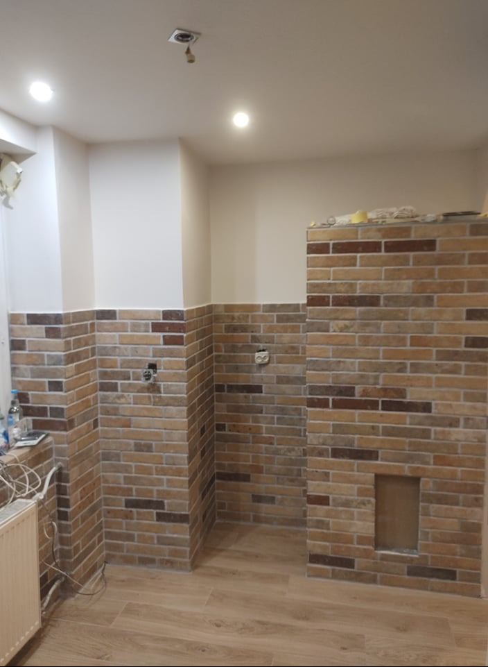 Częściowo wykończona kuchnia z ceglaną ścianą do połowy wysokości i drewnianą podłogą. Widoczne gniazdka elektryczne i fragment grzejnika.