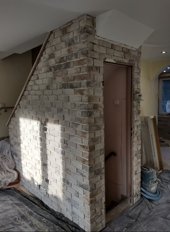 Wykończony fragment ściany z cegły dekoracyjnej nad drzwiami wewnątrz budynku. Widoczne narzędzia i zabezpieczenia podłogi. Nietypowe ułożenie cegły.