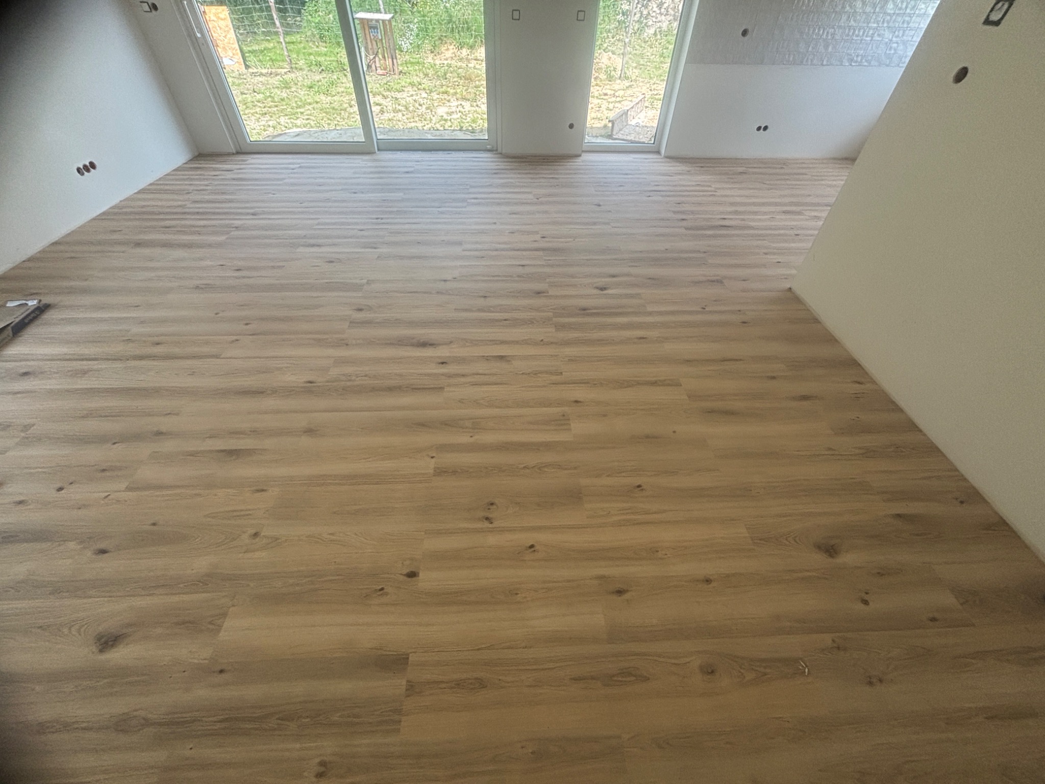 Jasna, drewnopodobna wykładzina w pokoju z oknami balkonowymi i skośnymi ścianami. Widoczne gniazdka elektryczne i fragment opakowania po wykładzinie.