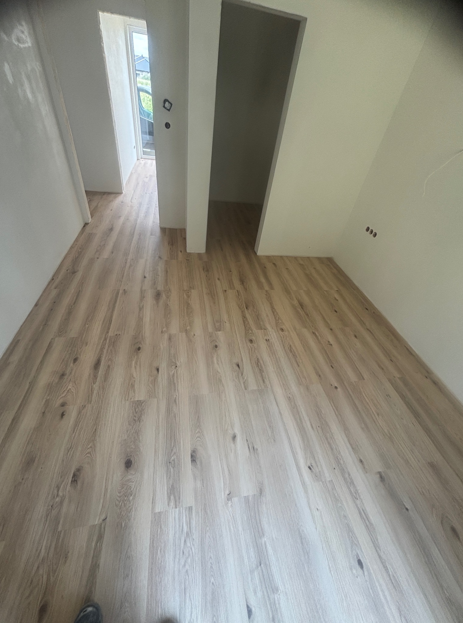 Jasna, drewnopodobna podłoga z paneli w świeżo wykończonym wnętrzu. Widoczne gniazdka elektryczne i fragment buta w lewym dolnym rogu. Dwa otwory drzwiowe.