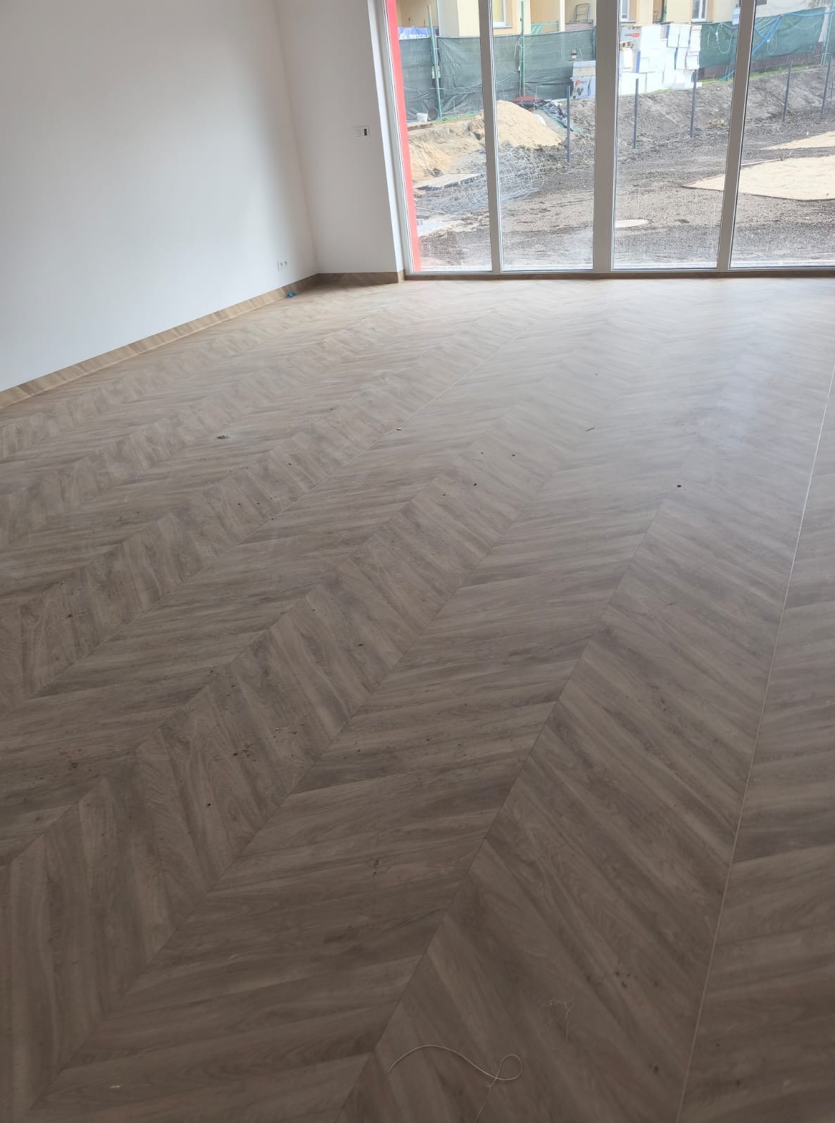 Ukończona podłoga z paneli ułożonych we wzór jodełki w jasnym, pustym pomieszczeniu z dużymi oknami. Widok na zewnątrz na plac budowy.