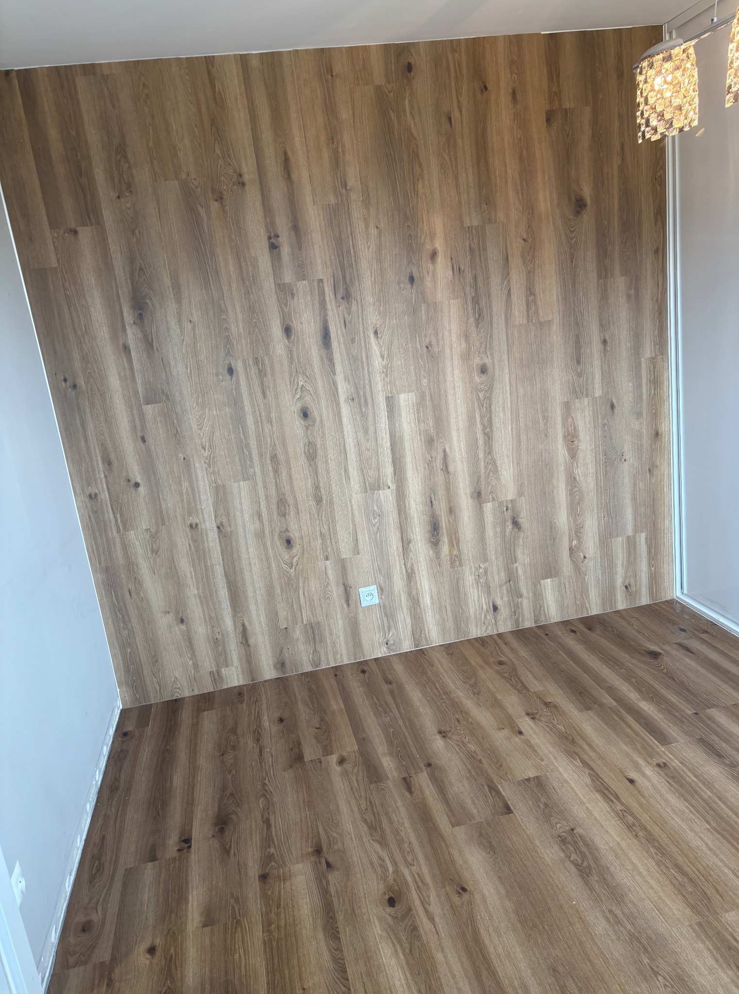 Wykończone wnętrze z drewnopodobną okładziną ścienną i podłogową, widoczny fragment lampy sufitowej. Całość w ciepłej tonacji, z gniazdkiem elektrycznym na ścianie.