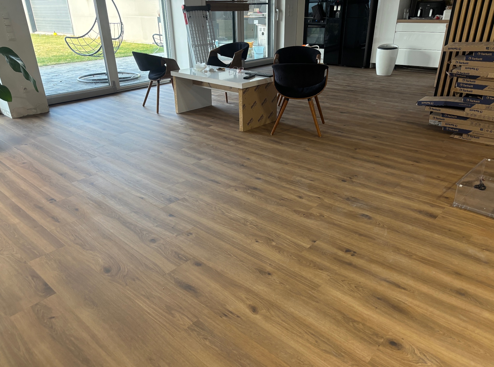 Ukończona podłoga z paneli w odcieniach brązu, z widocznymi paczkami paneli Tarkett. W tle okno z widokiem na ogród, krzesła i stolik.