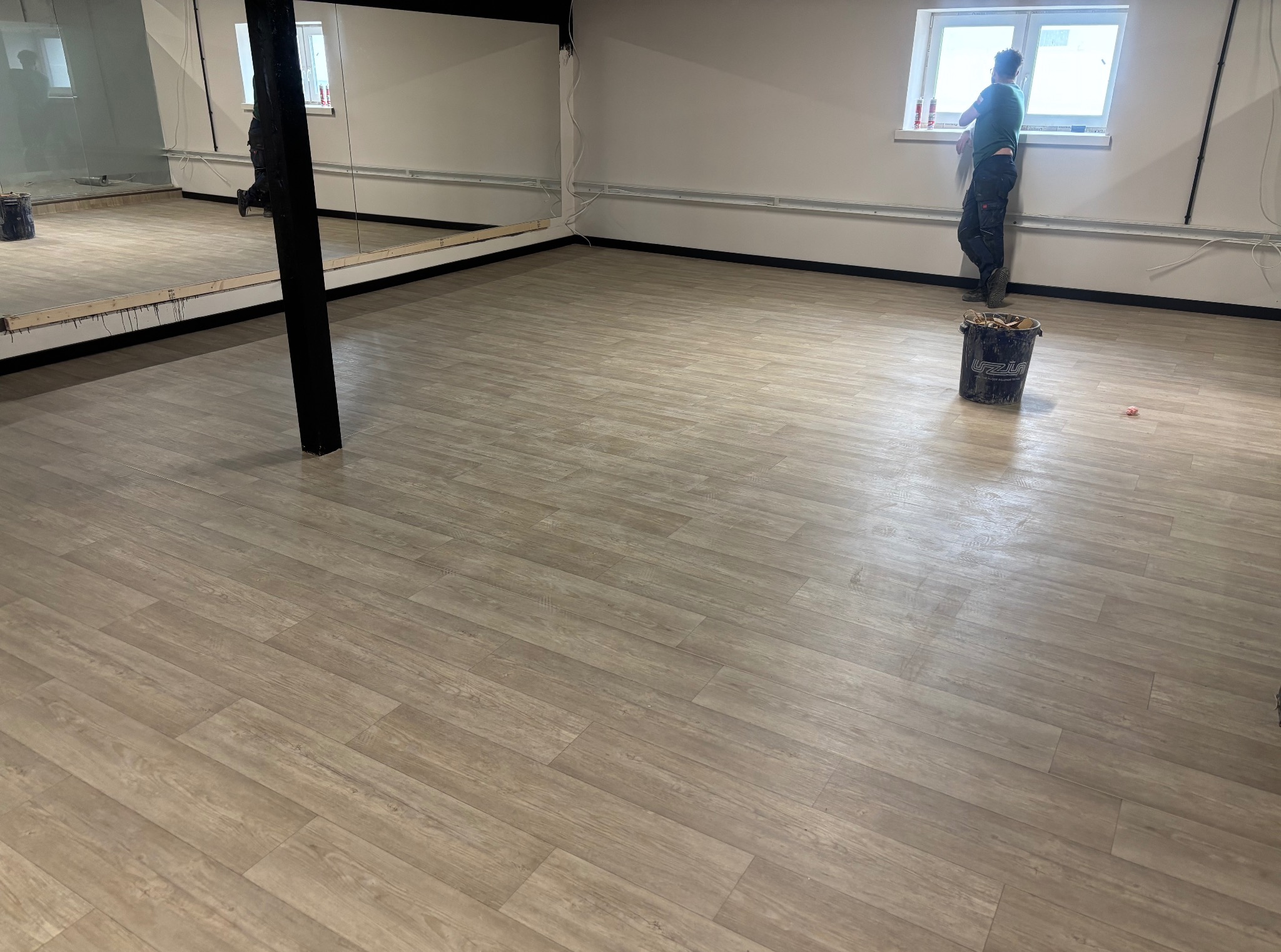 Jasna, drewnopodobna wykładzina w przestronnym wnętrzu z lustrem i oknem. Widoczny pracownik i wiadro z resztkami, sugerujące etap końcowy prac.