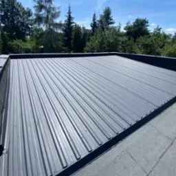 Szary dach z blachy trapezowej, widok z góry, na tle zieleni drzew.  Blacha przymocowana do dachu za pomocą wkrętów. Fragment dachu pokryty papą.