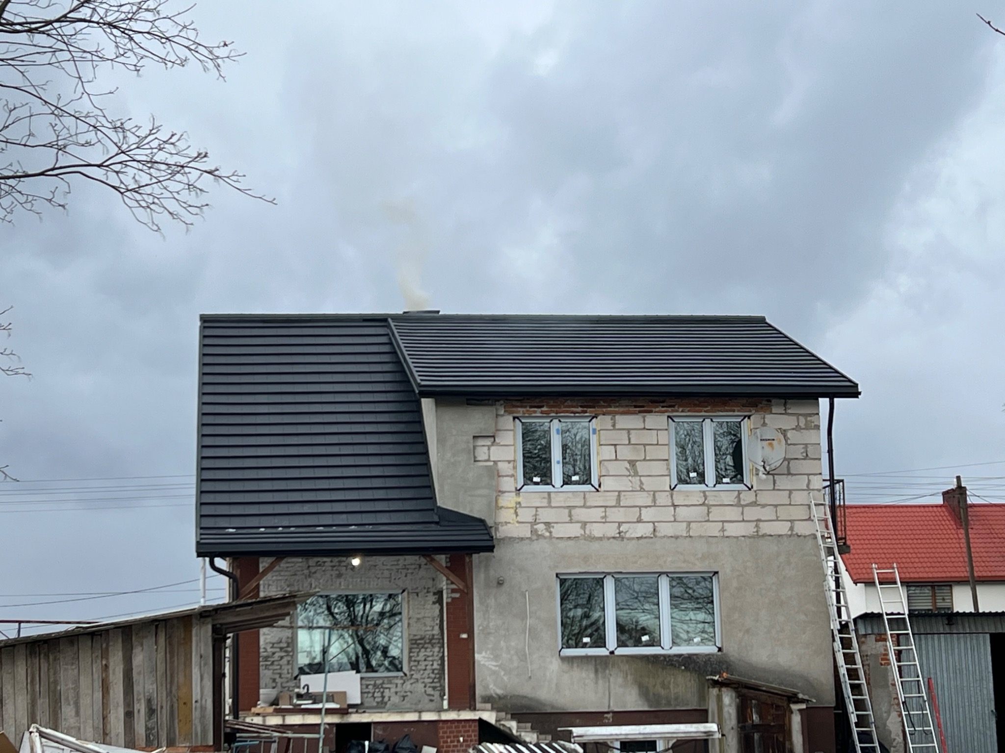 Dom w trakcie budowy z nowym, ciemnoszarym dachem z dachówki ceramicznej. Widoczne rusztowania i drabiny. Z komina unosi się dym.