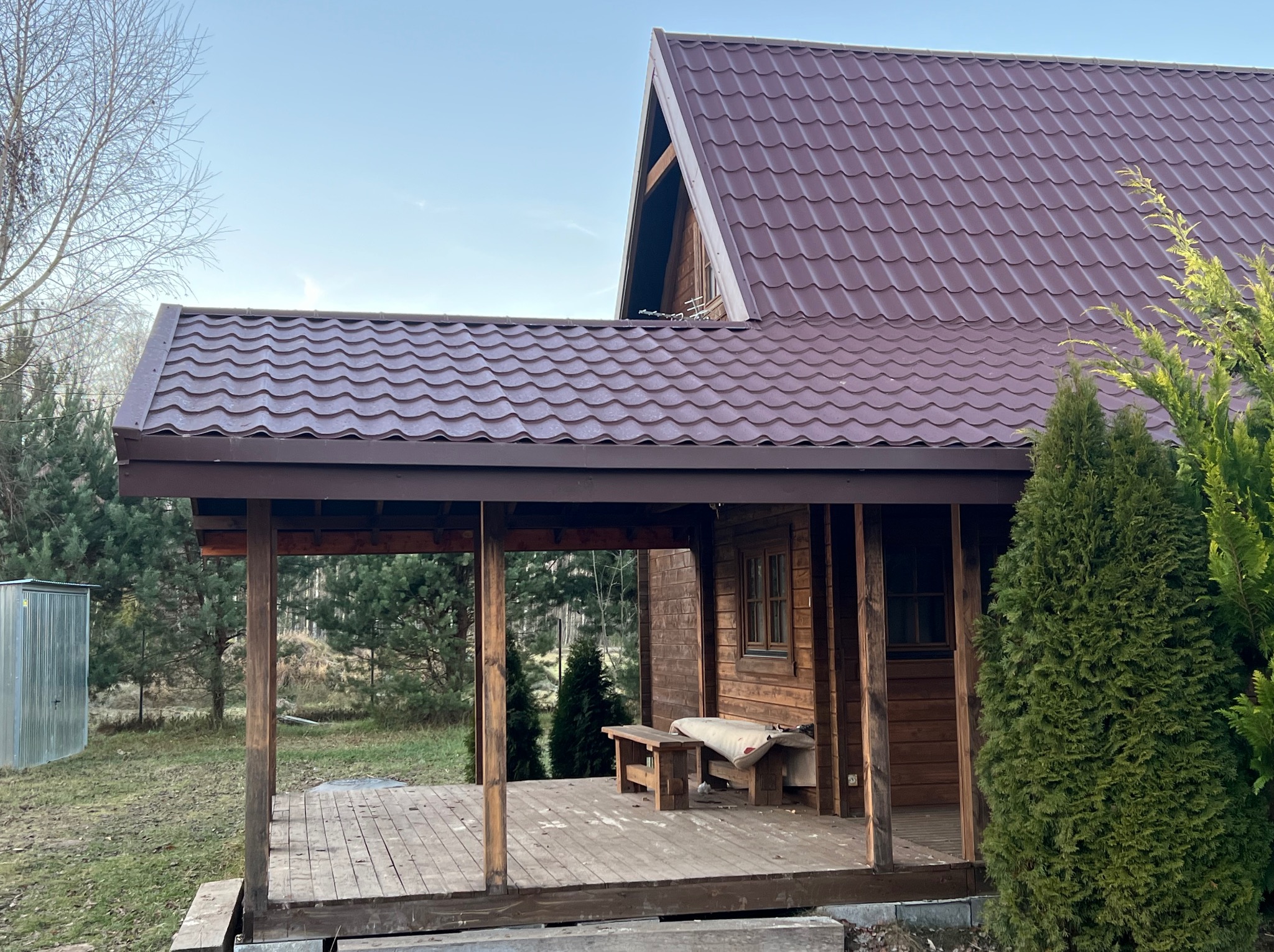 Dom z brązowym dachem z blachy trapezowej, widoczny z perspektywy werandy. Na dachu widoczna antena.