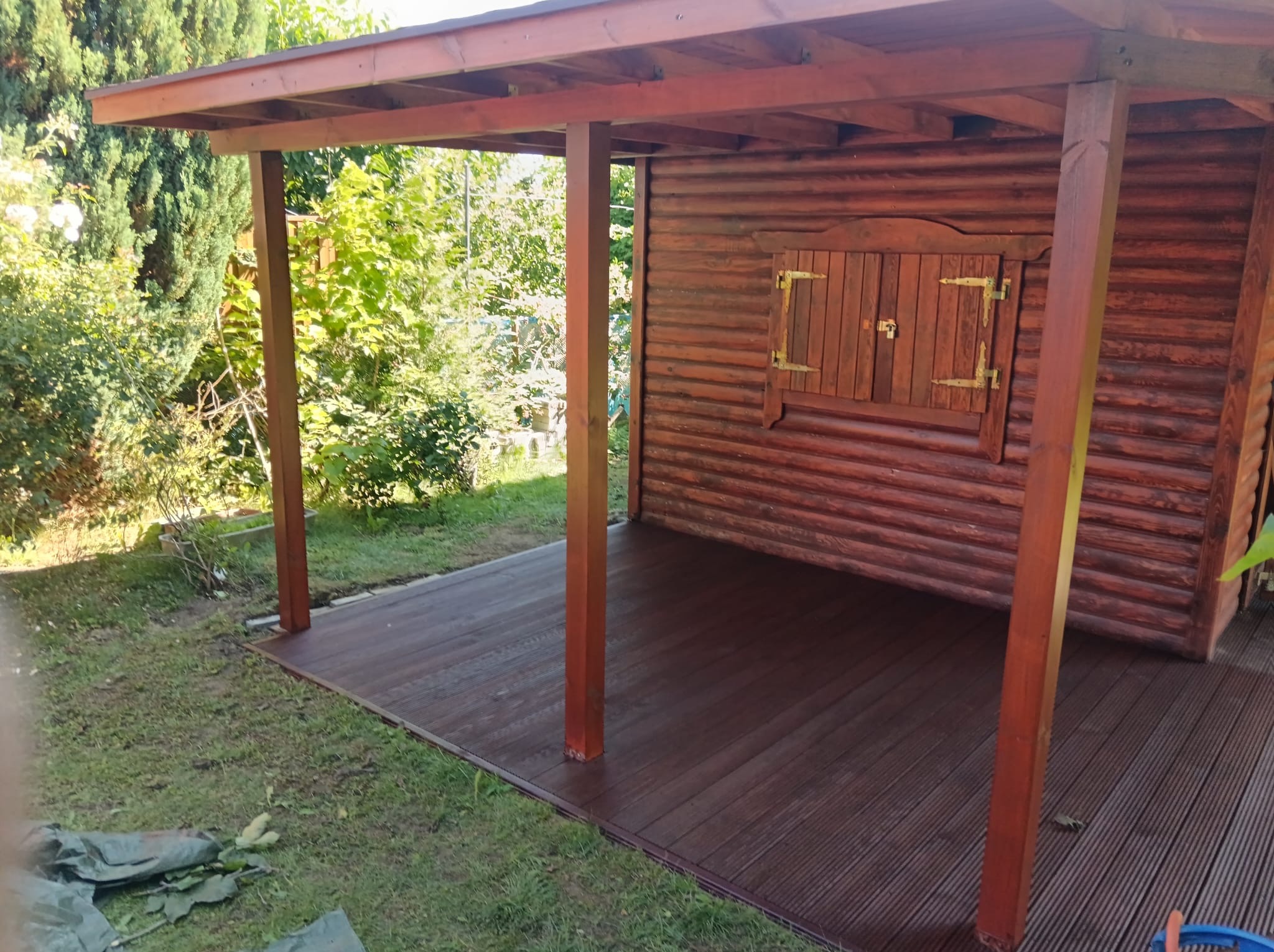 Drewniana altana z zamykanym oknem, świeżo zaimpregnowana, otoczona zielenią. Widoczna drewniana podłoga i konstrukcja dachu.