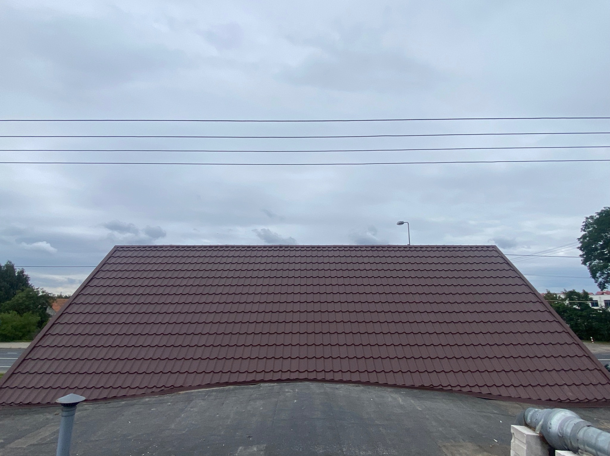 Brązowy dach z blachodachówki modułowej, widoczny z góry na tle pochmurnego nieba i linii energetycznych. Ujęcie podkreśla symetryczny układ paneli.