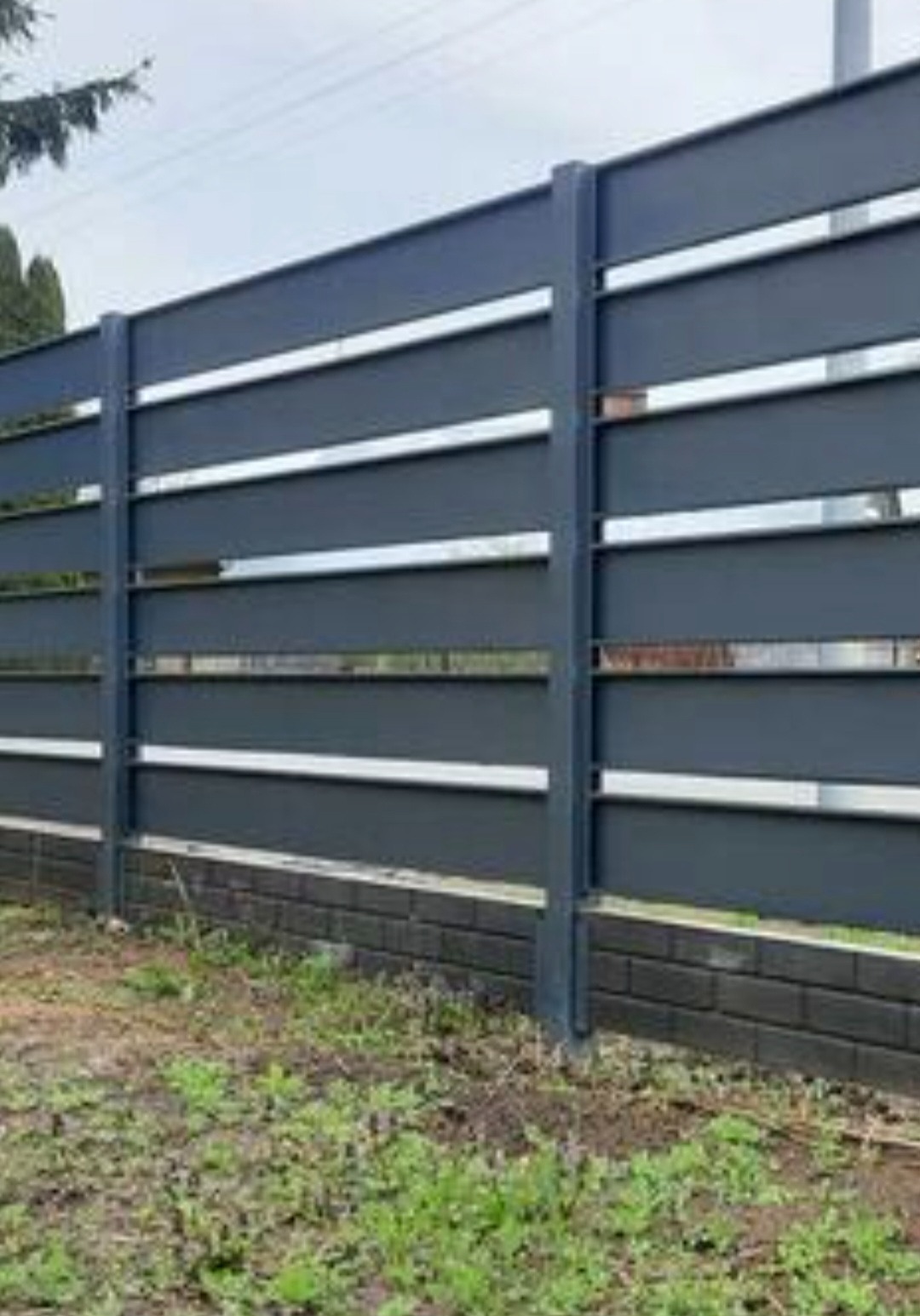Nowoczesne, grafitowe ogrodzenie panelowe z poziomymi, srebrnymi akcentami, osadzone na niskiej, ceglanej podmurówce. Widok z poziomu gruntu, trawa w tle.