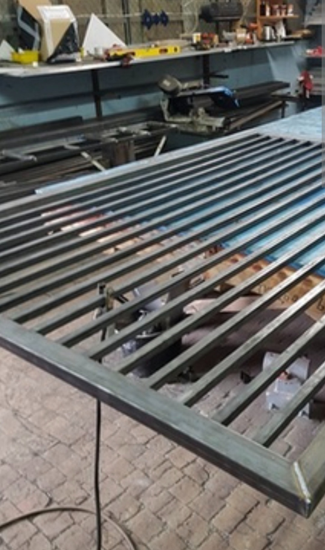 Surowa, spawana brama wjazdowa z metalowych profili, widoczna w warsztacie na tle narzędzi i materiałów. Szlifowane spawy, przygotowanie do malowania.