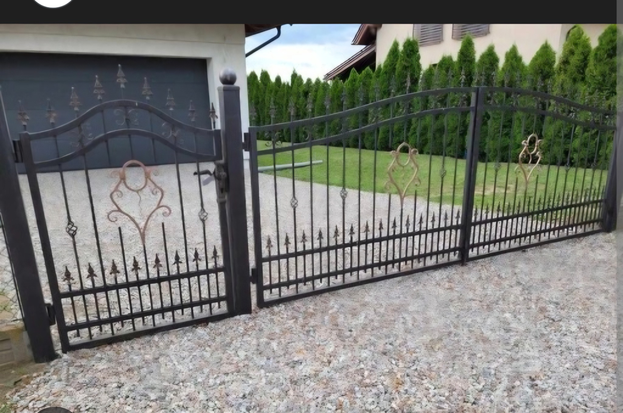 Czarna, metalowa brama wjazdowa z ozdobnymi elementami w kolorze miedzianym, na tle zielonych tuj i szarego podjazdu z kamienia. Widoczny fragment garażu.