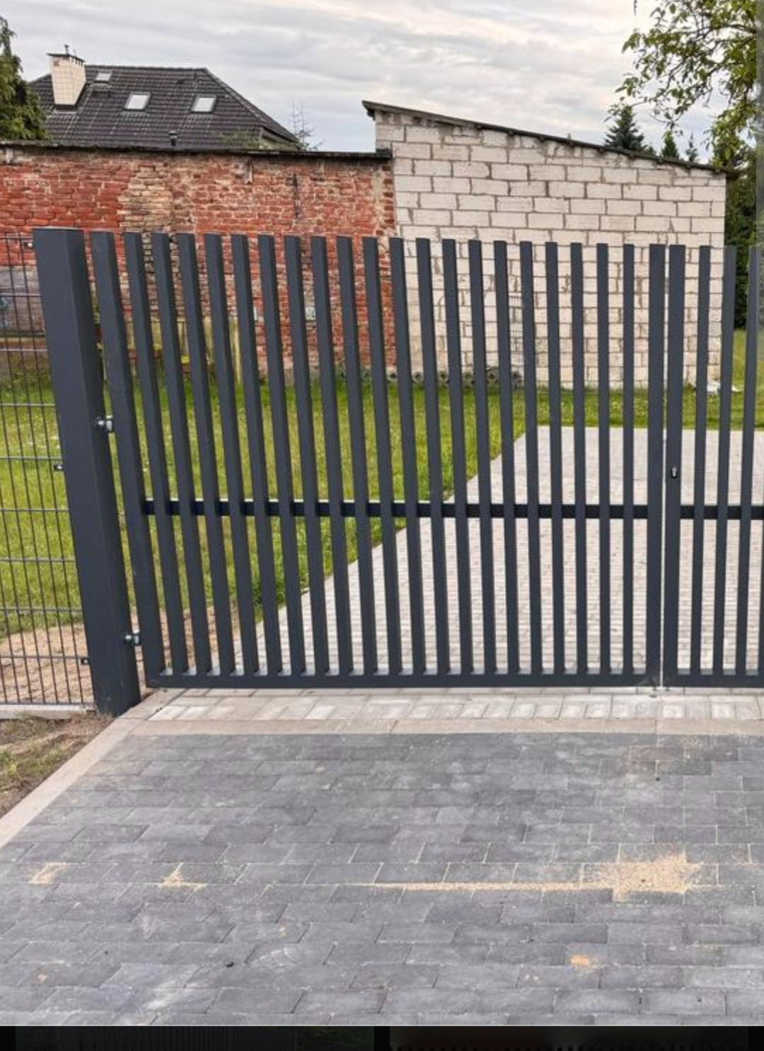 Nowoczesna, grafitowa brama wjazdowa z pionowych profili, widok z bliska na tle muru z cegły i kostki brukowej. Minimalistyczny design.