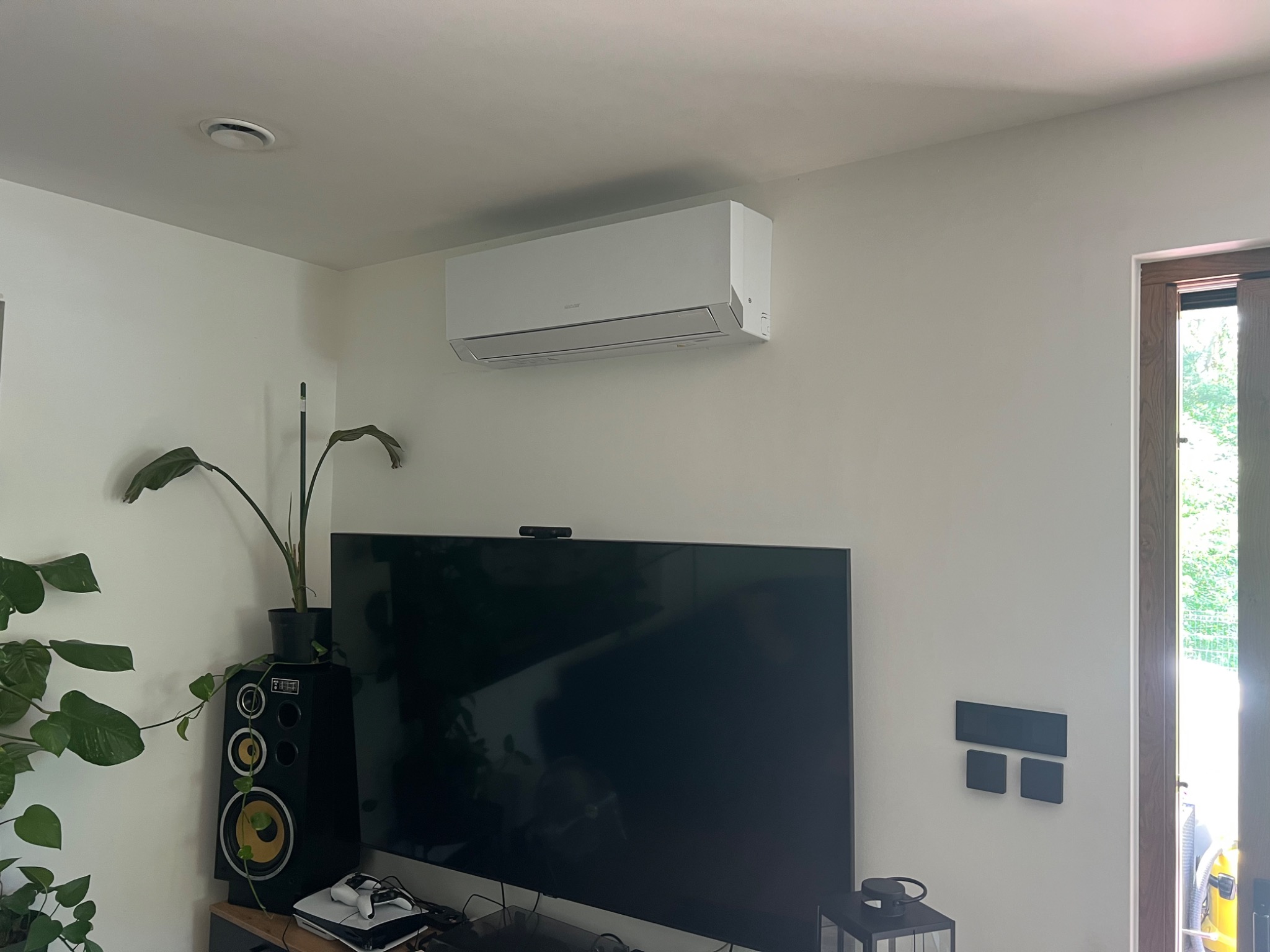 Biała klimatyzacja ścienna w salonie, nad telewizorem. Widoczny głośnik i roślina doniczkowa. Minimalistyczne wnętrze z drewnianymi drzwiami balkonowymi.