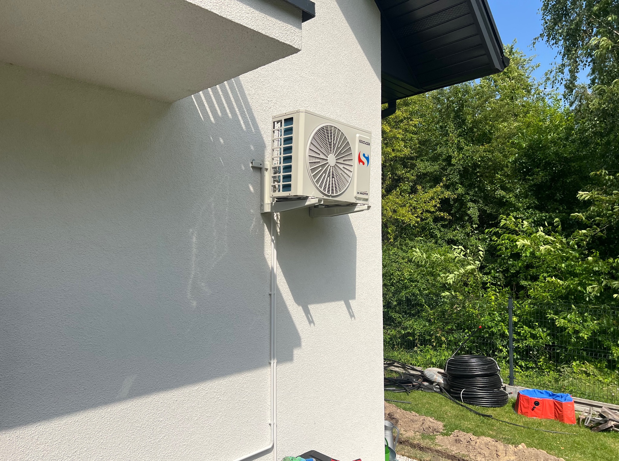 Biała jednostka zewnętrzna klimatyzacji Sinclair zamontowana na ścianie budynku z jasną elewacją. W tle zieleń drzew i krzewów, widoczne elementy instalacji na trawniku.