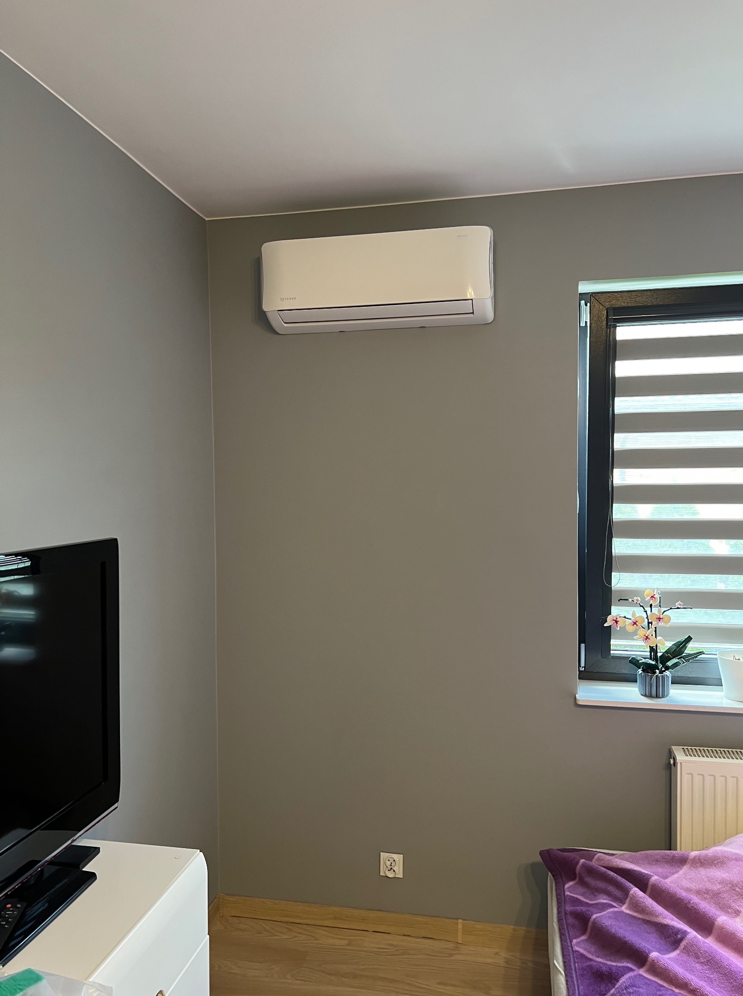Biały klimatyzator ścienny marki Totenso zamontowany w szarym pokoju z oknem i roletami. Widoczny fragment telewizora i łóżka. Minimalistyczny design.