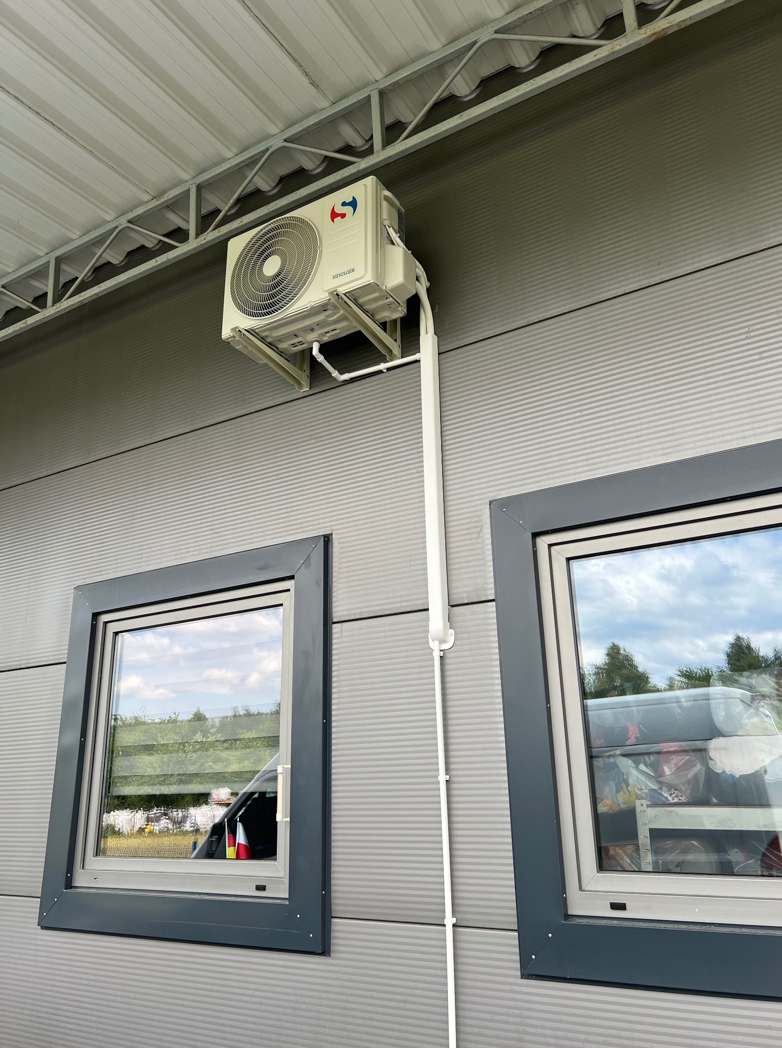 Zamontowany klimatyzator Sinclair na szarej elewacji budynku z dwoma oknami. Biała rura odprowadzająca skropliny. Widok z dołu na urządzenie.