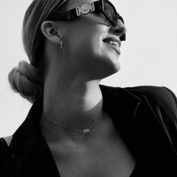 Nel Maczkiewicz - Czarno-białe ujęcie kobiety w okularach i marynarce, spoglądającej w górę. Widoczny profil, delikatny uśmiech, biżuteria: kolczyki i naszyjnik. Minimalistyczny styl.