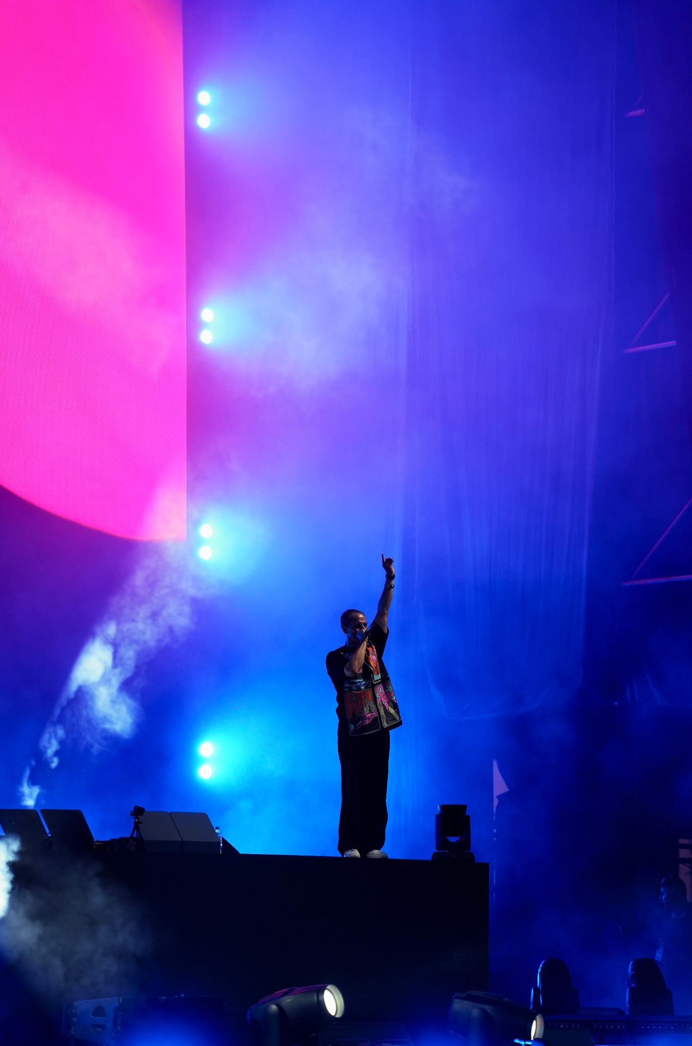 Piosenkarz na scenie w intensywnym oświetleniu, z mikrofonem w ręku i uniesioną dłonią, występuje na tle różowo-niebieskich świateł i sprzętu scenicznego.