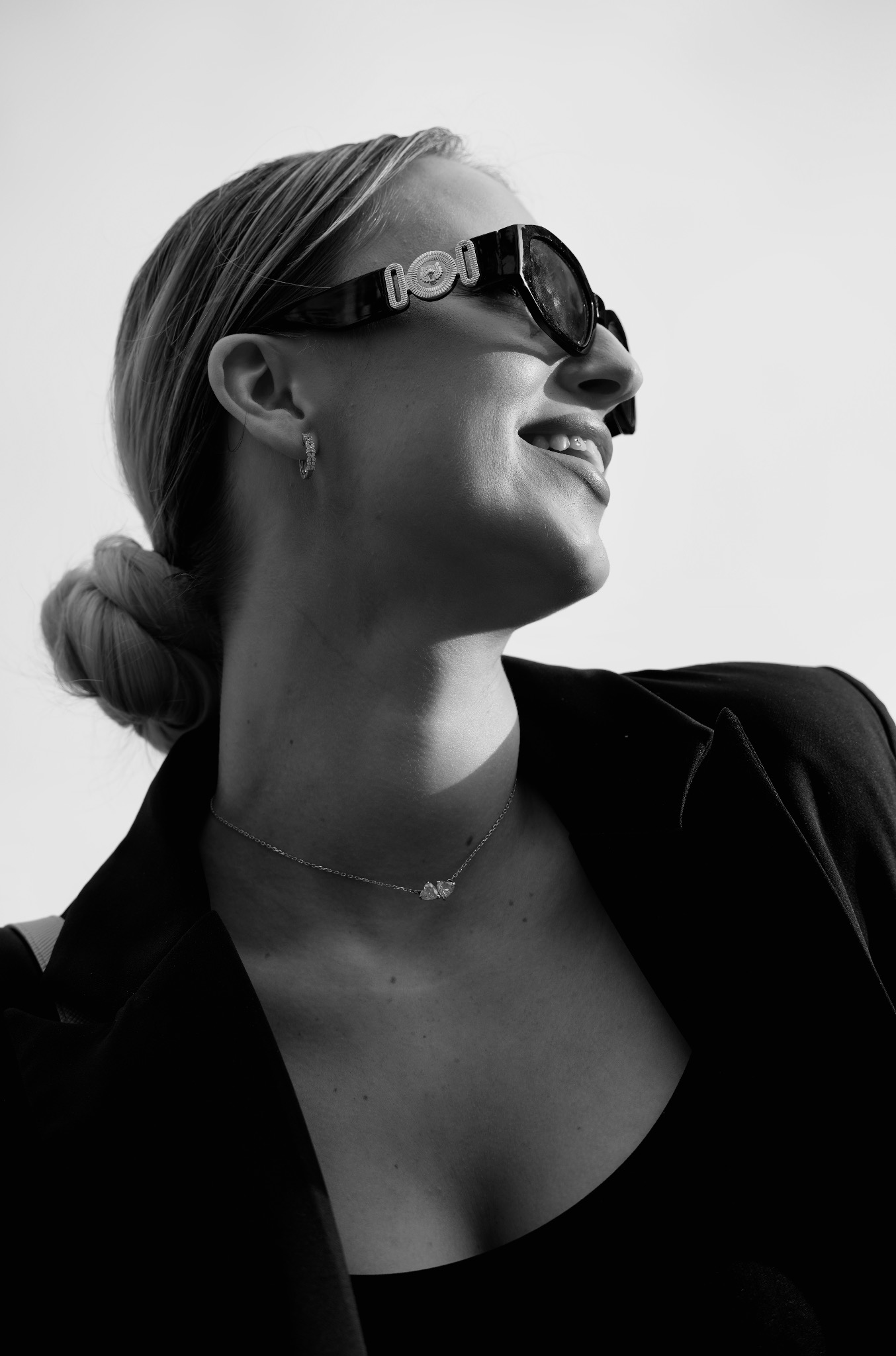 Czarno-białe ujęcie kobiety w okularach i marynarce, spoglądającej w górę. Widoczny profil, delikatny uśmiech, biżuteria: kolczyki i naszyjnik. Minimalistyczny styl.