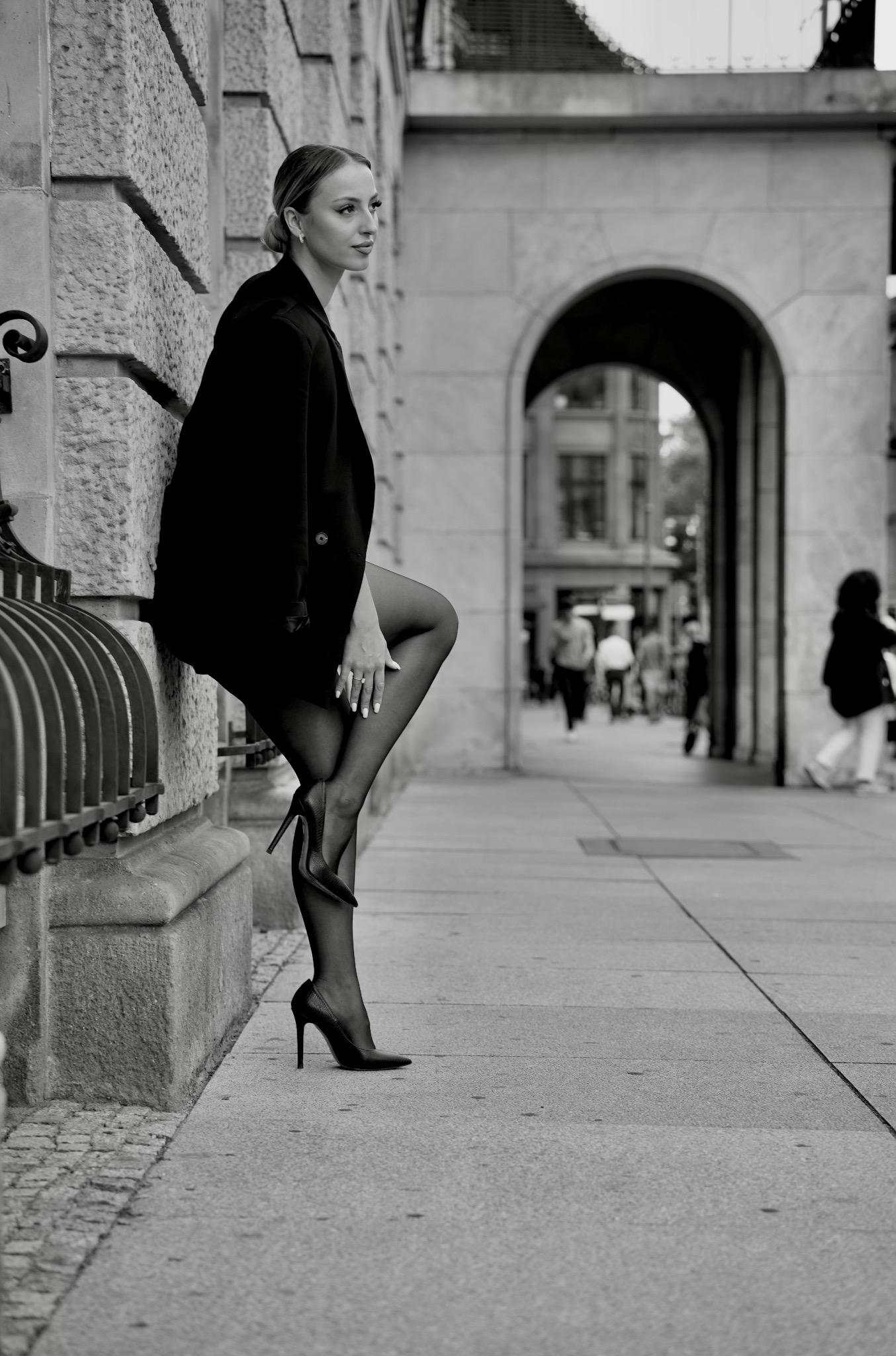 Czarno-białe zdjęcie kobiety w czarnym żakiecie i szpilkach, opierającej się o kamienną ścianę na tle miejskiej architektury z przejściem w formie łuku.