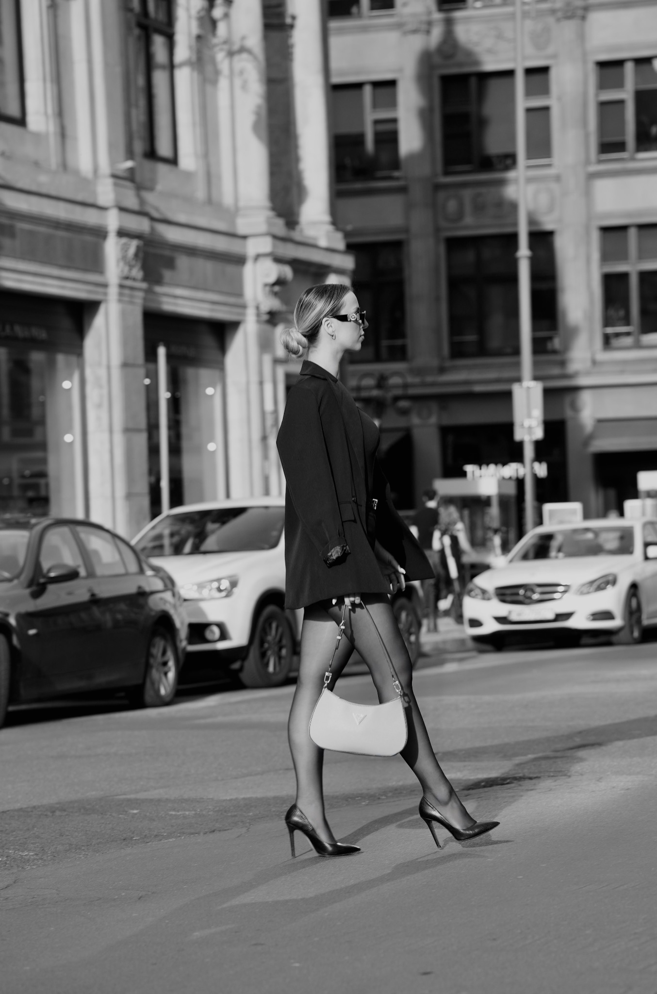 Czarno-białe zdjęcie kobiety w modnej stylizacji przechodzącej przez ulicę. Ubrana w marynarkę, rajstopy i szpilki, trzyma torebkę. W tle budynki i samochody.