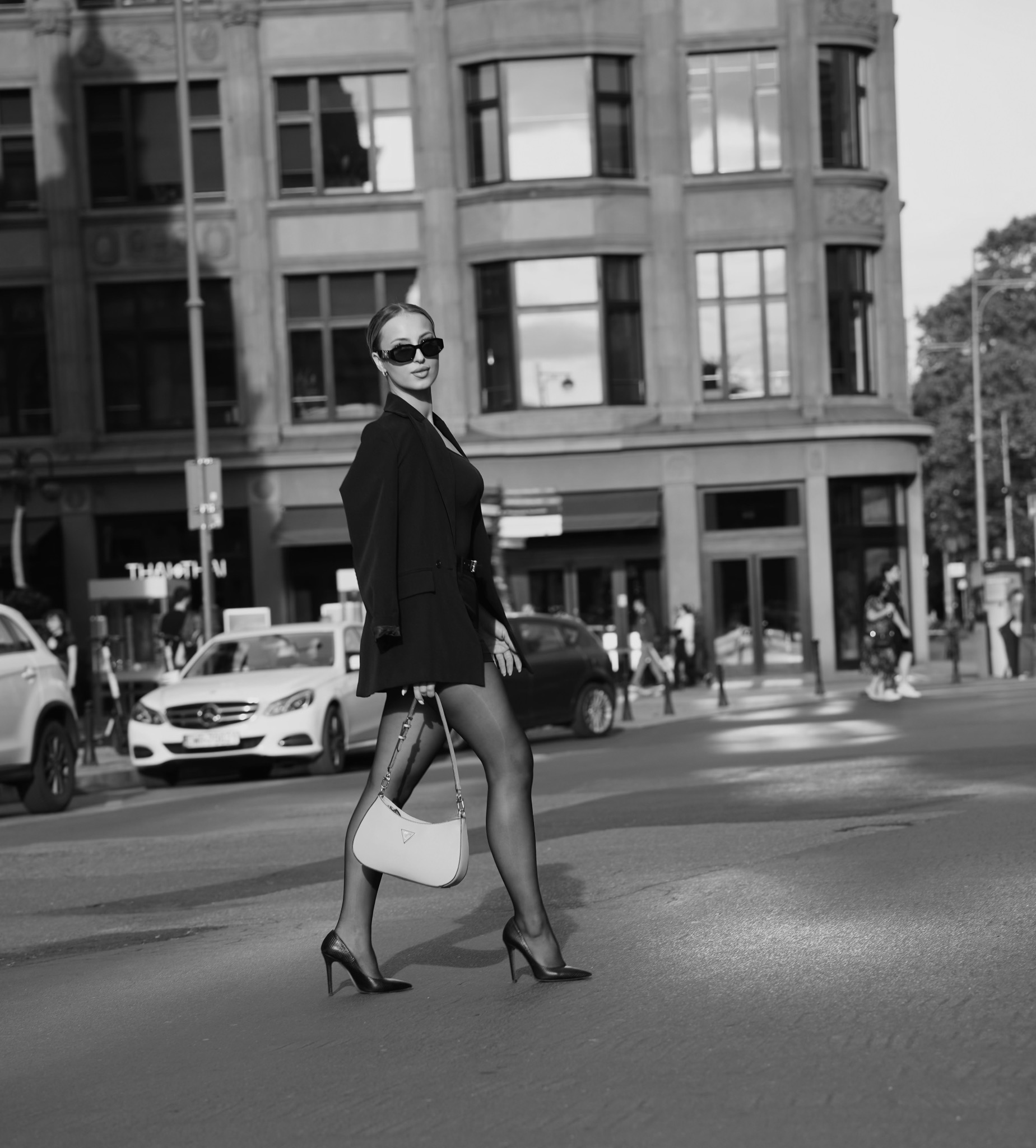 Stylowa kobieta w czarnym garniturze i okularach przechodzi przez ulicę na tle miejskiej zabudowy. Uwagę przykuwają rajstopy i szpilki oraz torebka na ramieniu.