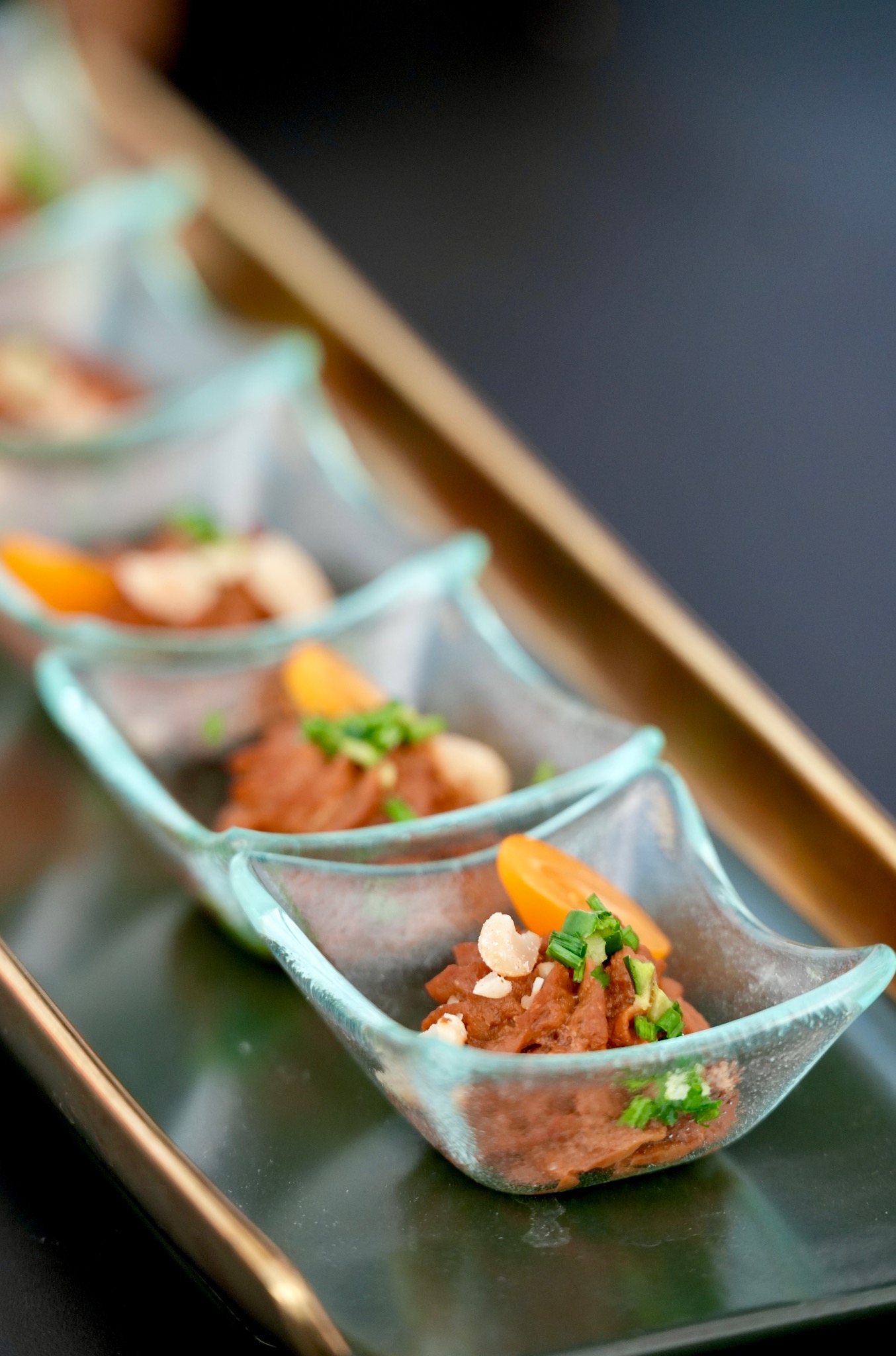 Elegancko podane przystawki w szklanych naczyniach, udekorowane zieleniną i pomarańczowymi elementami, ustawione na ciemnym, błyszczącym talerzu z złotą ramką.