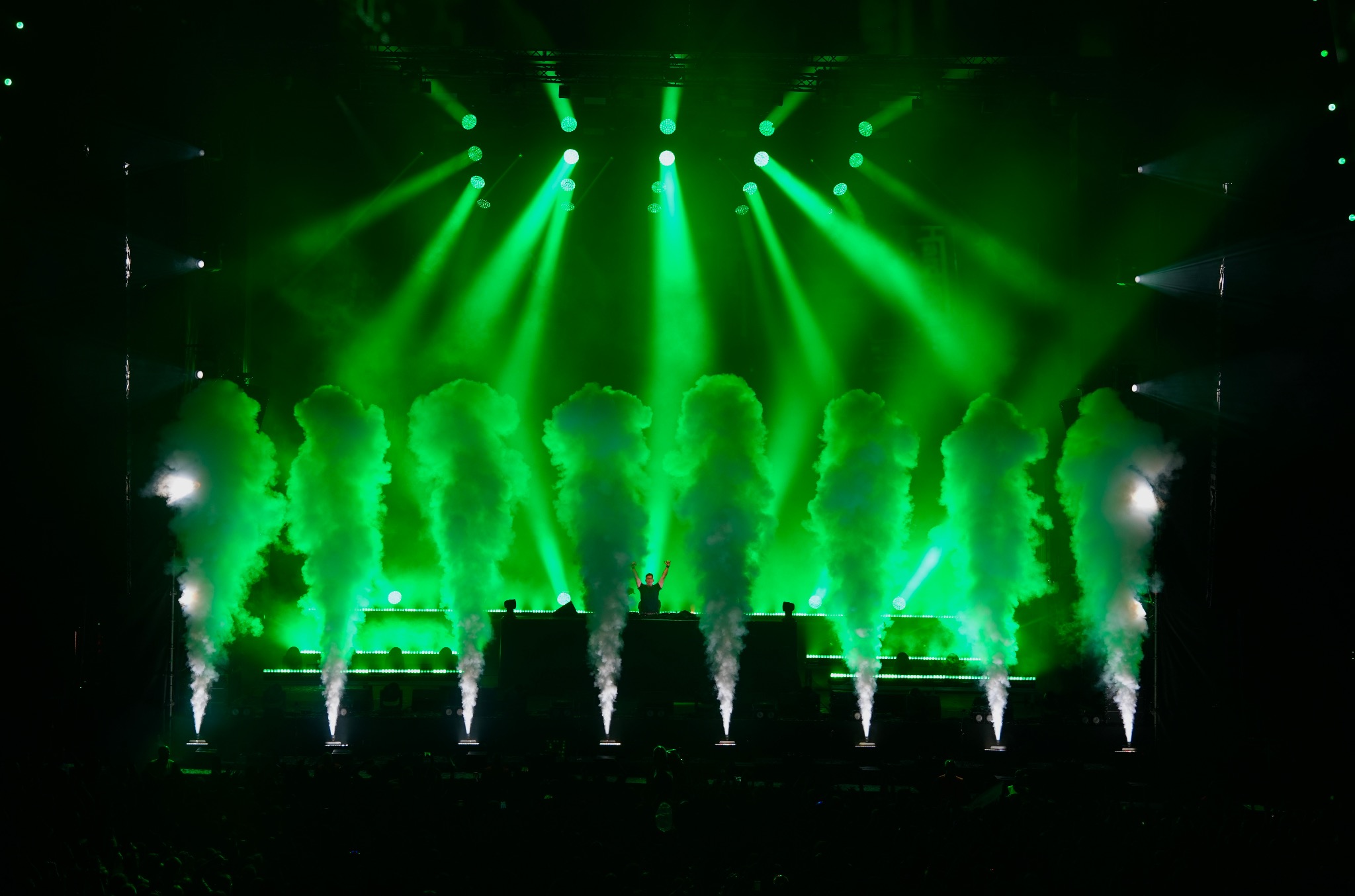 DJ na scenie podczas eventu, otoczony zielonymi światłami i dymem. Widoczny tłum publiczności. Dynamiczne ujęcie z koncertu.