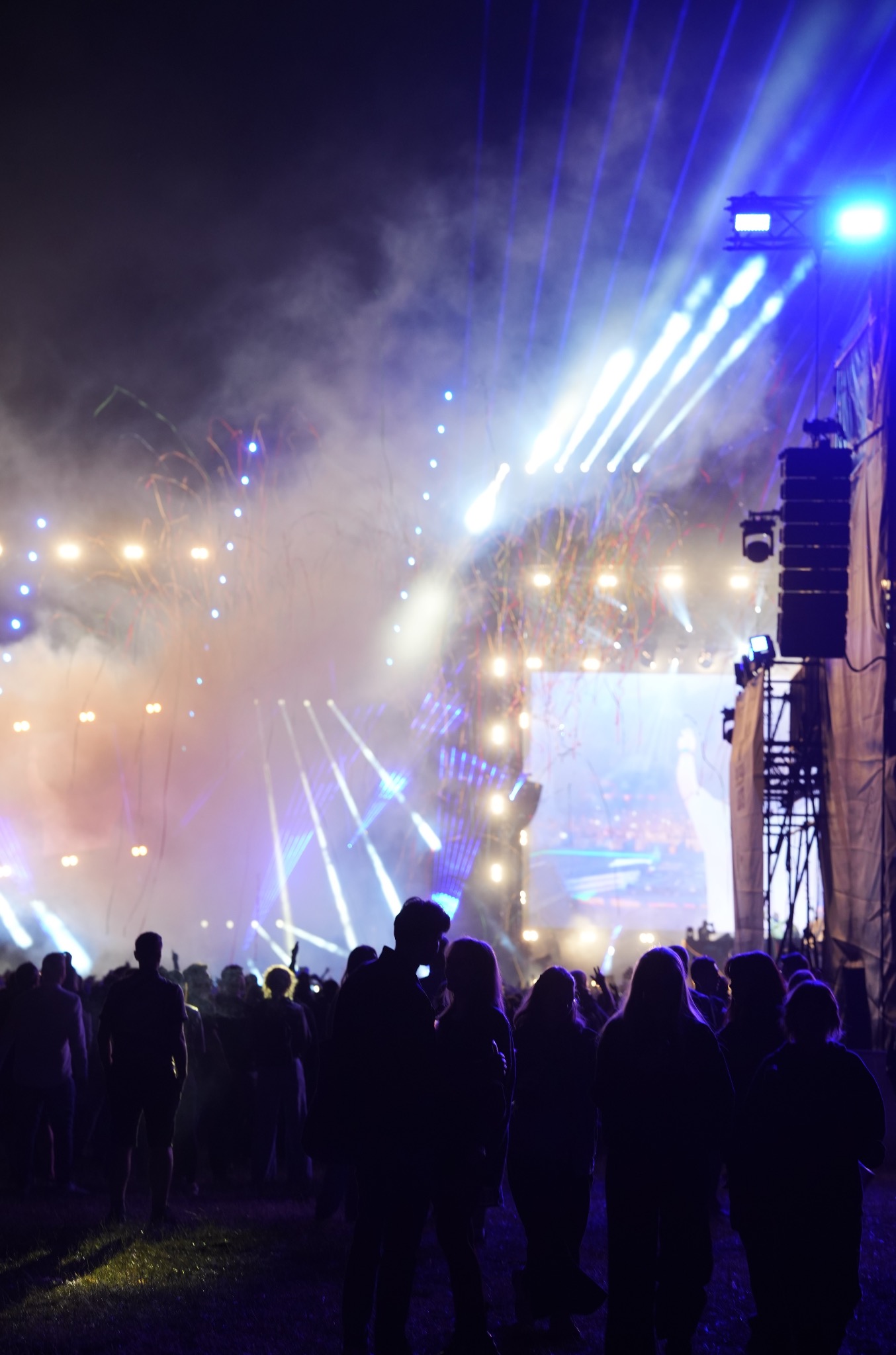 Tłum ludzi w ciemności obserwuje scenę z efektami świetlnymi i konfetti podczas koncertu plenerowego. Widoczne sylwetki publiczności i oświetlenie sceniczne.