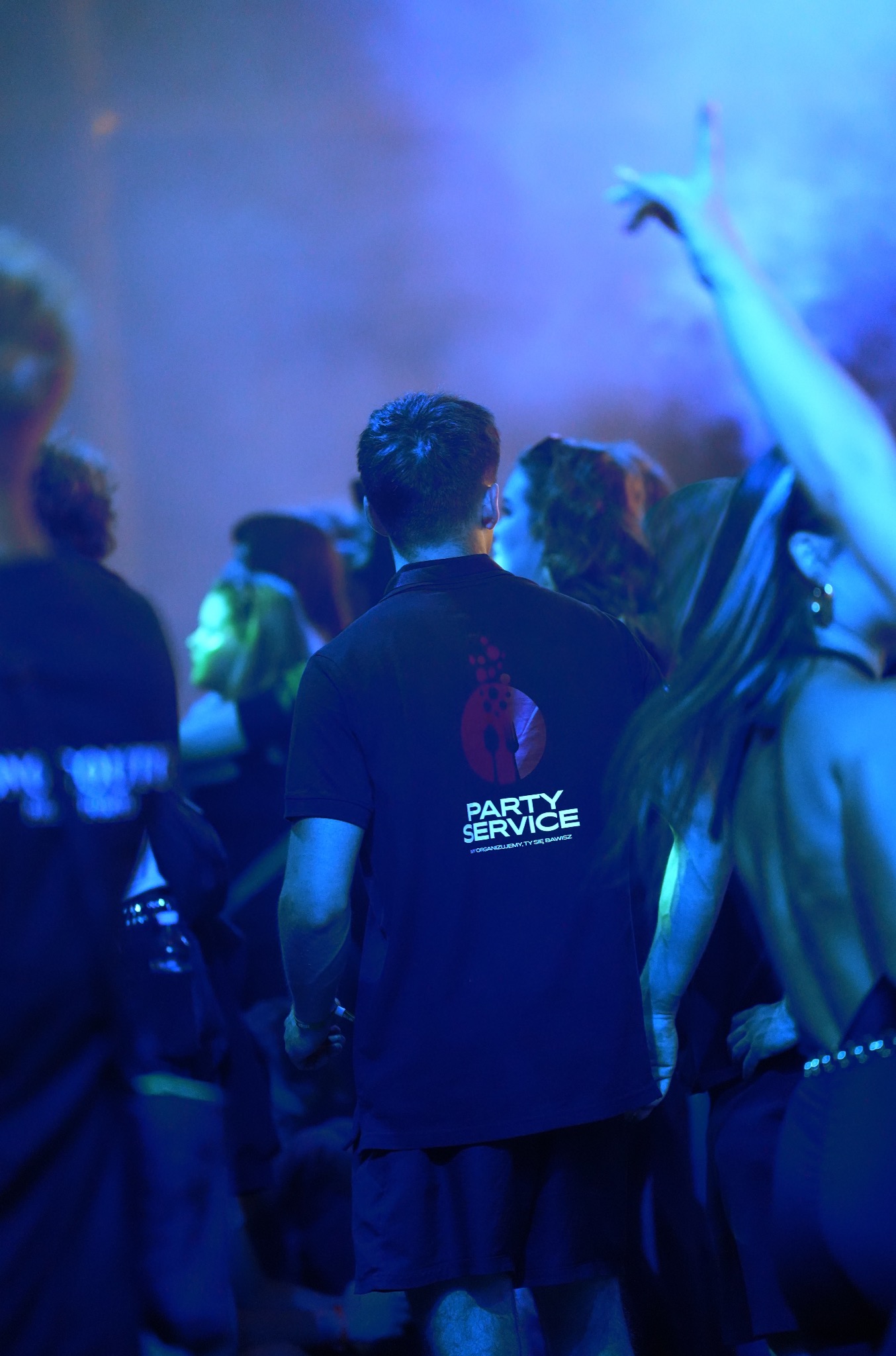 Pracownik w koszulce 'Party Service' na tle roztańczonego tłumu w niebieskim oświetleniu, sugerujący obsługę imprezy.